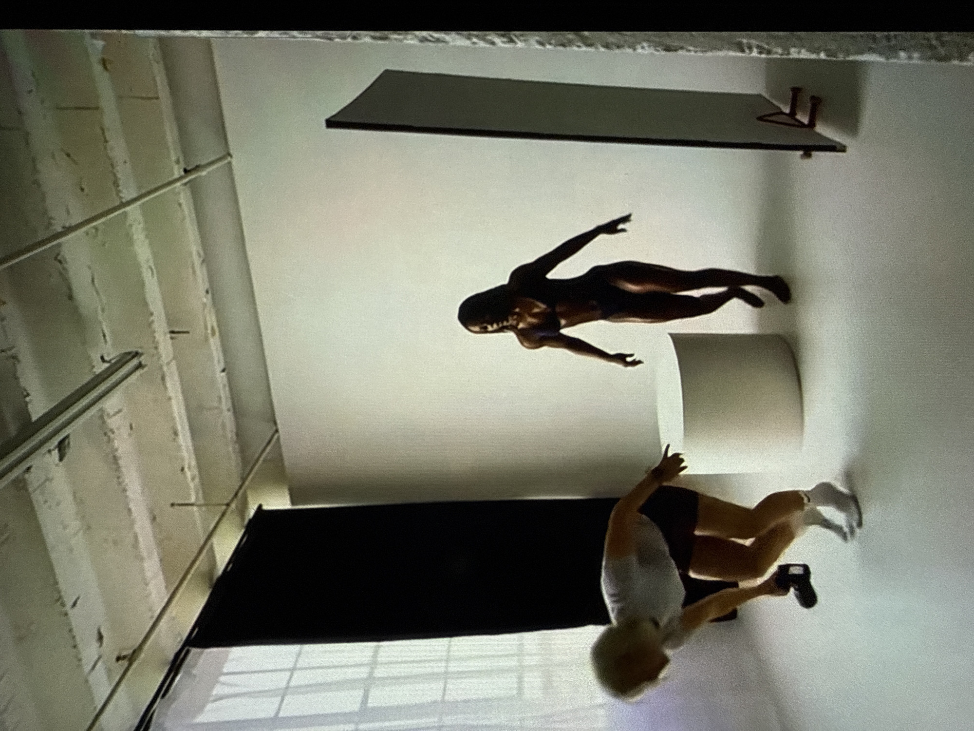The image depicts an indoor photography or video studio located in Varna, Bulgaria. The environment features a seamless white floor and wall cyclorama on the right side. On the left, a large window with a visible grid pattern is partially covered by a vertical black fabric backdrop or curtain. The ceiling reveals exposed concrete beams, metal rails, and utility pipes.
Two individuals are present. On the left, in the foreground, a person with short, light-colored hair, wearing a light-colored t-shirt, dark shorts, and white socks, is bent forward at the waist. They are holding a dark, camera-like device in their right hand, pointing it towards the floor.
In the mid-ground on the right, a second individual with dark skin and long dark hair, wearing a dark two-piece swimsuit, is standing upright with arms slightly bent and hands open, palms facing downwards. This individual is barefoot and appears to be in motion, possibly walking or posing.
Between the two individuals, a white cylindrical pedestal or prop is situated on the white floor. On the far right, a tall, dark rectangular panel, possibly a reflector or a divider, stands vertically with a small brown support structure at its base. The scene is illuminated by what appears to be a combination of natural light from the window and overhead ambient lighting.