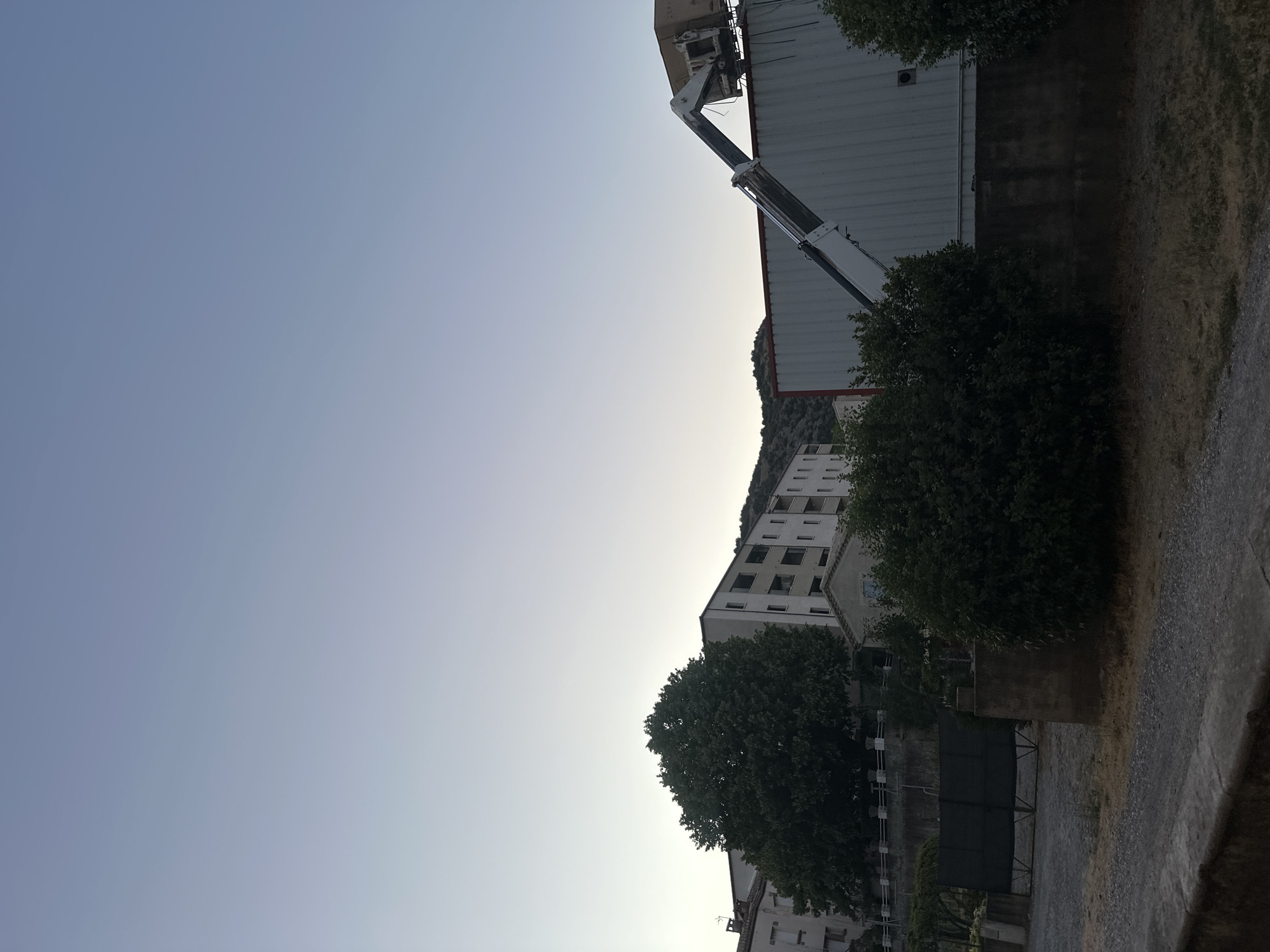 This image depicts a view of the city of Anduze, France, during what appears to be dusk or dawn, given the gradient of light in the sky. In the foreground, there is a gravel pathway leading towards a dark metal gate, flanked by lush green bushes on the right. Beyond the gate, a stone wall with a railing is visible, separating the foreground from a larger building complex.
Dominating the mid-ground are several multi-story buildings, characteristic of urban architecture. A particularly large, leafy tree stands prominently in front of one of these buildings. To the right, a corrugated metal structure is visible, topped with what appears to be a crane or lifting mechanism, with a light-colored box or container being held by its arm. Hills are visible in the background, suggesting the city's location within a more mountainous or hilly region. The overall atmosphere is tranquil, with the muted light and stillness of the scene.