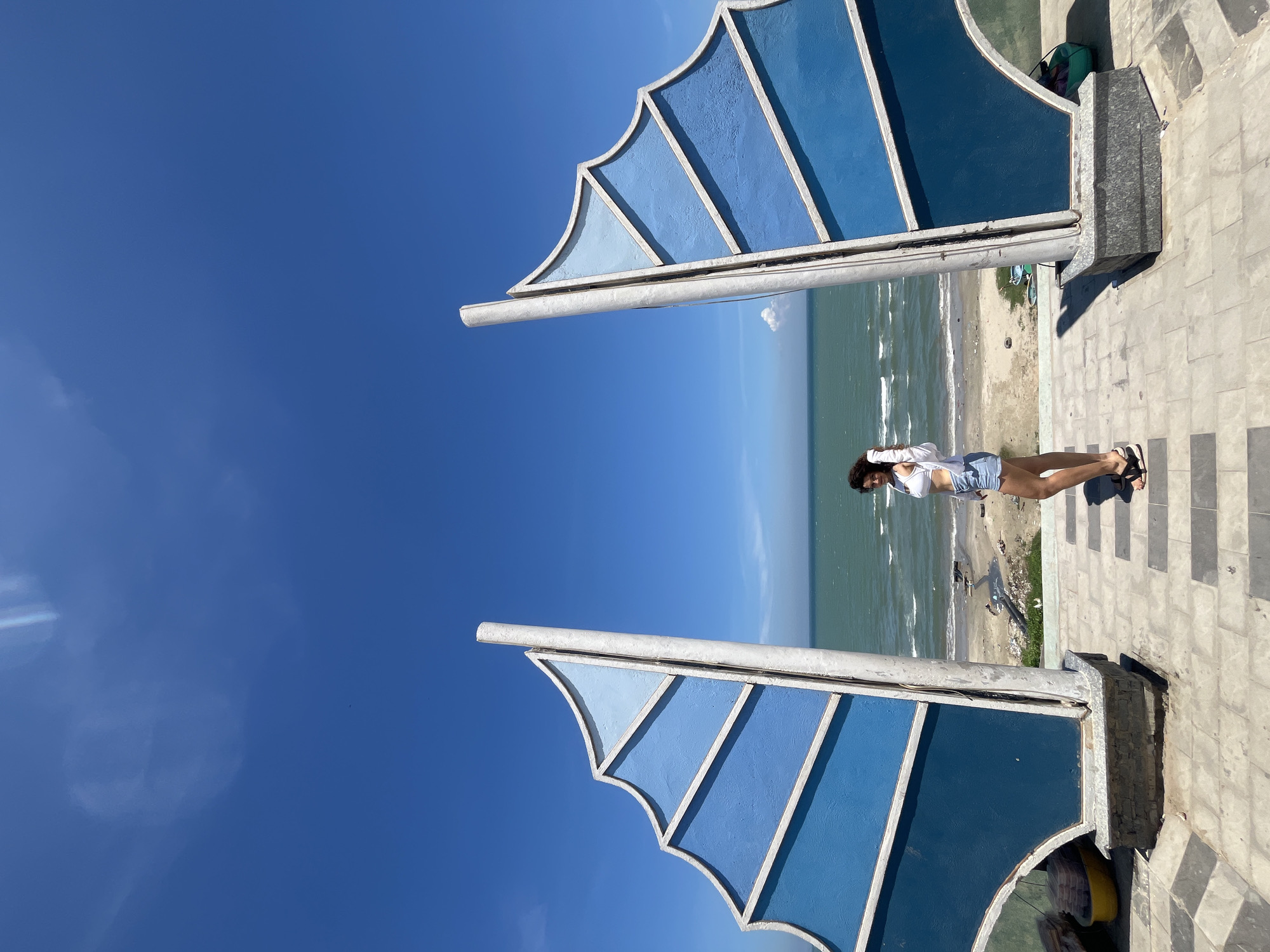 An image taken in Mũi Né, Vietnam, shows a young woman standing in an open pathway with the ocean in the background. The pathway is paved with gray and black square tiles. On either side of the pathway are large, blue, sail-like structures with white trim. The woman is positioned in the center of the frame, between these structures, and is facing slightly to her left. She is wearing a white crop top, denim shorts, and black sandals. Her left hand is raised, touching her hair. The ocean is visible beyond the structures, with waves rolling onto a sandy beach. The sky is clear and bright blue with a few wispy clouds. The overall scene suggests a sunny day at a coastal location.