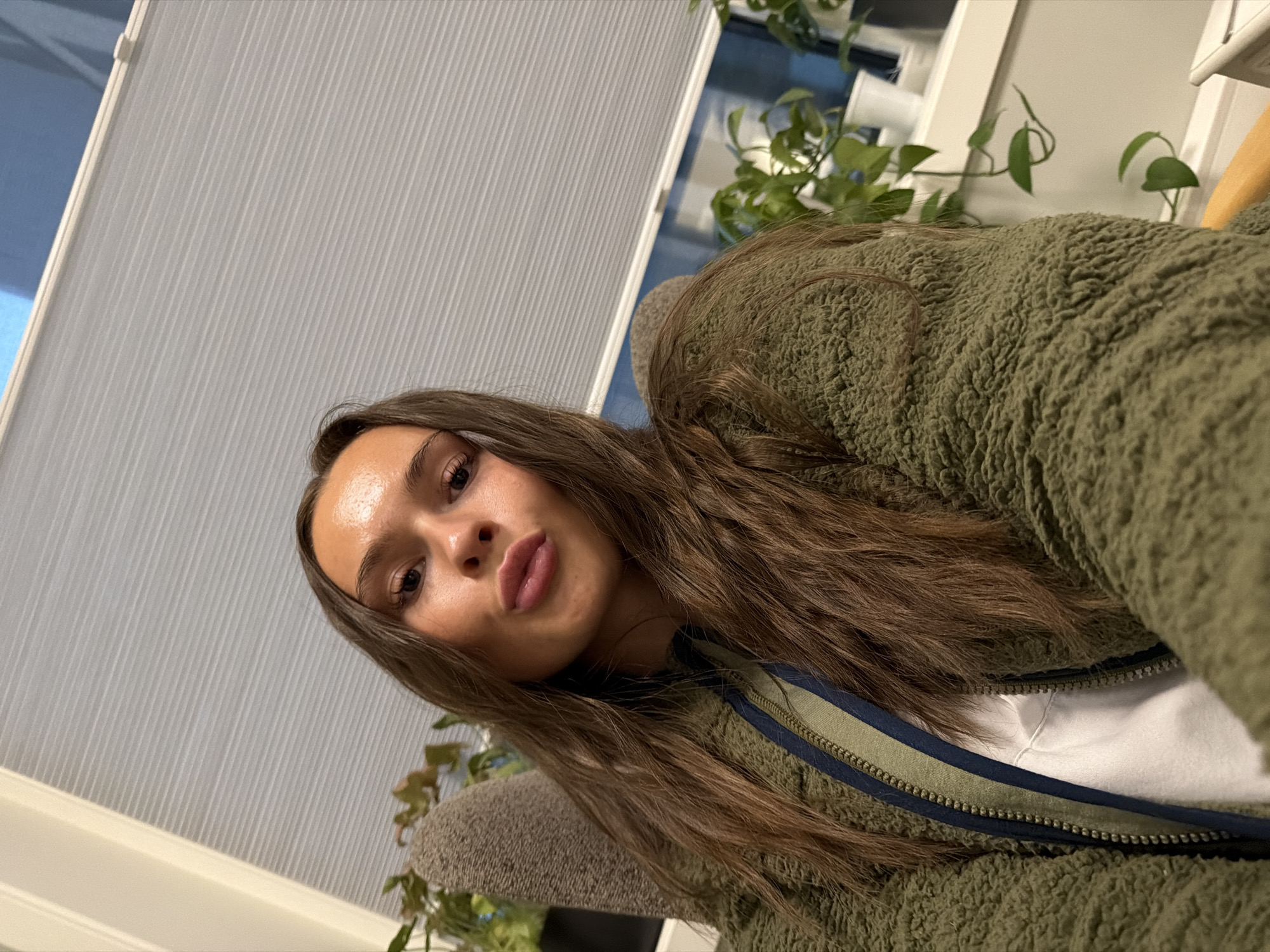 A young woman with long brown hair is taking a selfie in a room. She is wearing a green, fuzzy jacket over a white shirt. Her lips are pursed. She is sitting in a chair, and to her left is a gray, textured surface, possibly an armchair. Behind her is a window covered by horizontal blinds. On the windowsill, there are several potted plants. The lighting in the room is somewhat dim. The location is the city of Harstad, Norway.
