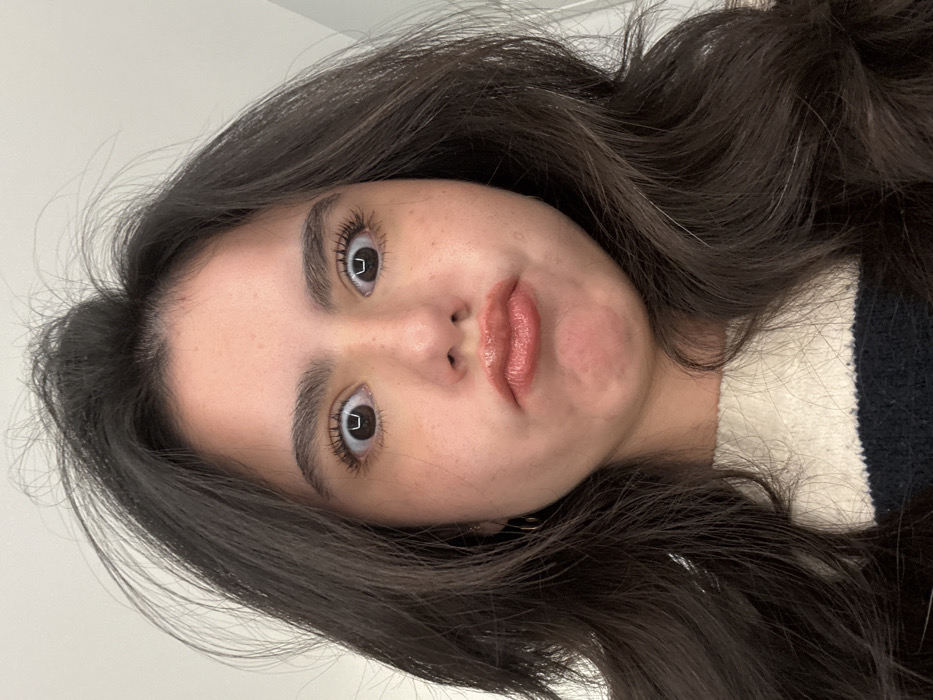 A young woman tilts her head, facing the camera. She has fair skin, dark brown wavy hair, and dark eyebrows. Her eyes are wide, and she has full lips with a glossy finish. She wears a dark sweater with a white collar. The background is a plain white wall. Her expression is slightly comical, with her lips pursed.