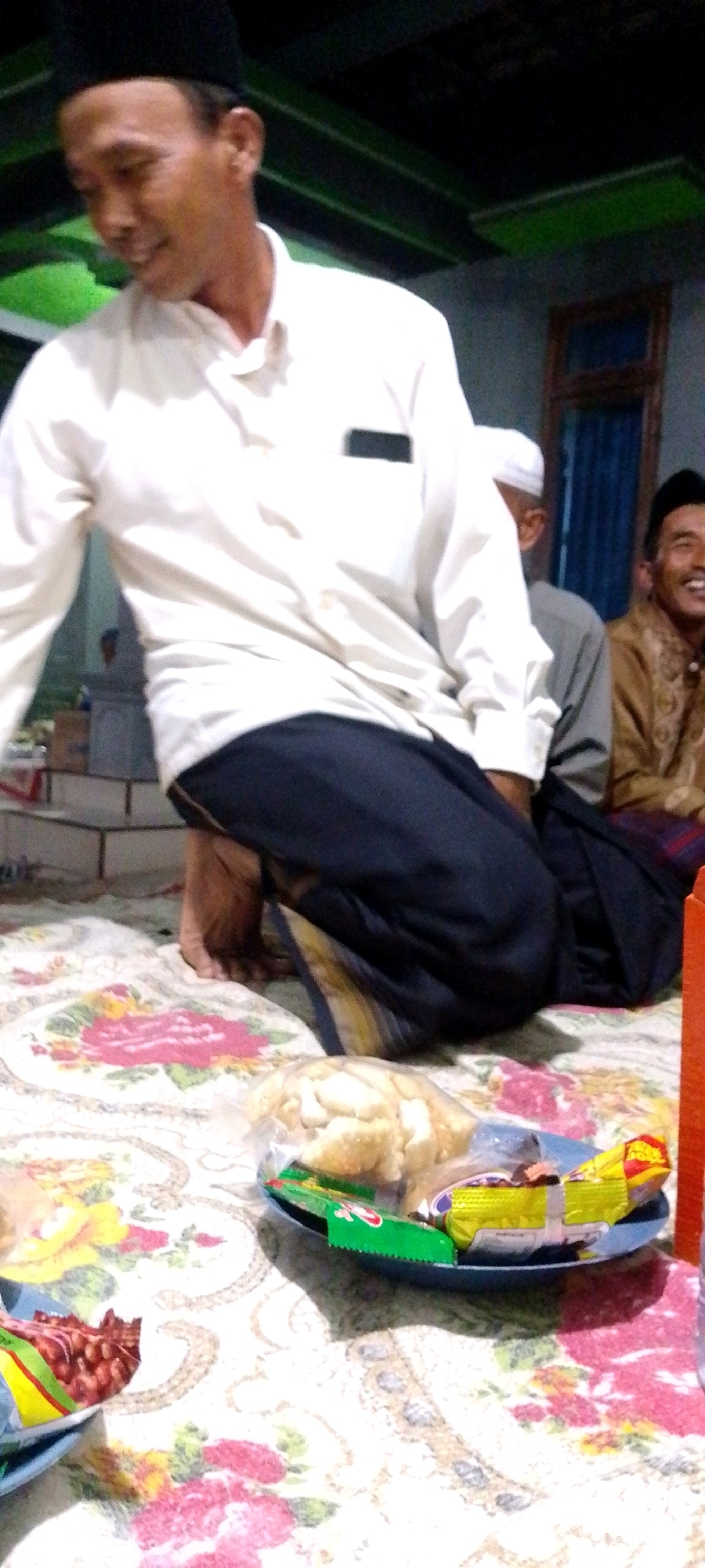 A group of men are gathered indoors. The man in the foreground is wearing a white long-sleeved shirt, a black peci hat, and dark pants. He is looking down and smiling. To his right, another man in a white peci hat and grey shirt is visible. Further to the right, a man in a gold patterned garment is laughing. They are seated on a patterned rug, with a plate of snacks and food in front of them. The environment appears to be a home or community gathering space.