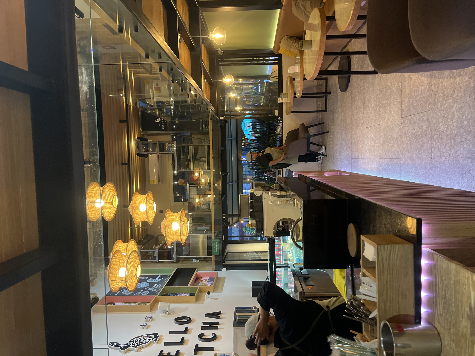 This photograph shows the interior of a cafe or restaurant. The location is identified as San Bernardino Tlaxcalancingo, Mexico. A cafe worker is behind the counter, visible partially in the foreground. Several woven lamps provide light. Through a window, there are patrons. The cafe is clean, well-lit, and features a mix of textures. The interior design includes wooden shelving and modern lighting fixtures.