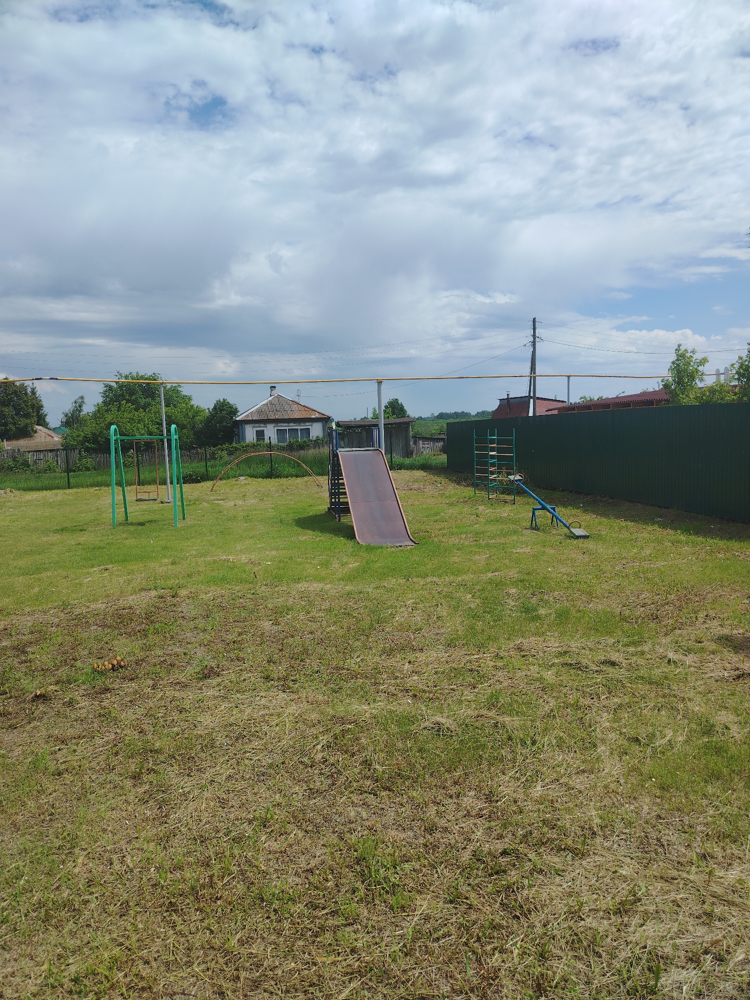 The image depicts an outdoor scene in Klyuchi, Russia, likely a residential area. The foreground is dominated by a patch of mown grass, showing some dry, yellowed blades. In the mid-ground, a small playground is visible. This includes a red metal slide, a green metal swing set with a single swing, and a blue seesaw. Behind the playground, a dark green corrugated metal fence runs along the right side of the image. Beyond the fence, trees and residential buildings are partially visible, including a white house with a grey roof. The background features a cloudy sky with patches of blue. The overall impression is of a simple, rural or suburban outdoor space.