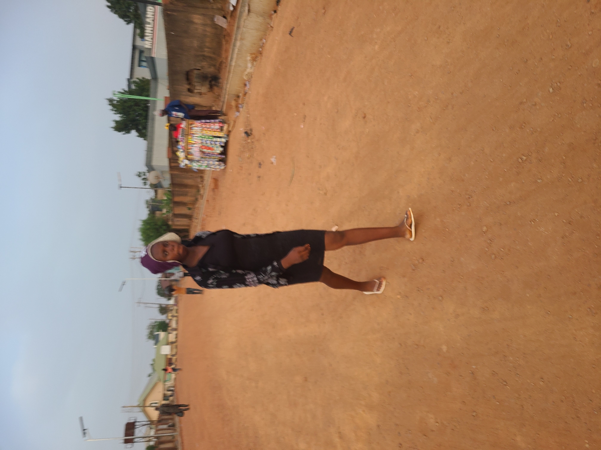 A woman in a dark patterned dress and light sandals stands on a dirt road in Jos, Nigeria. She is wearing a head covering. Behind her, a vendor with a cart displays colorful items for sale. A building with the word "MAINLAND" visible is in the background, along with other buildings and trees. Utility poles line the road.