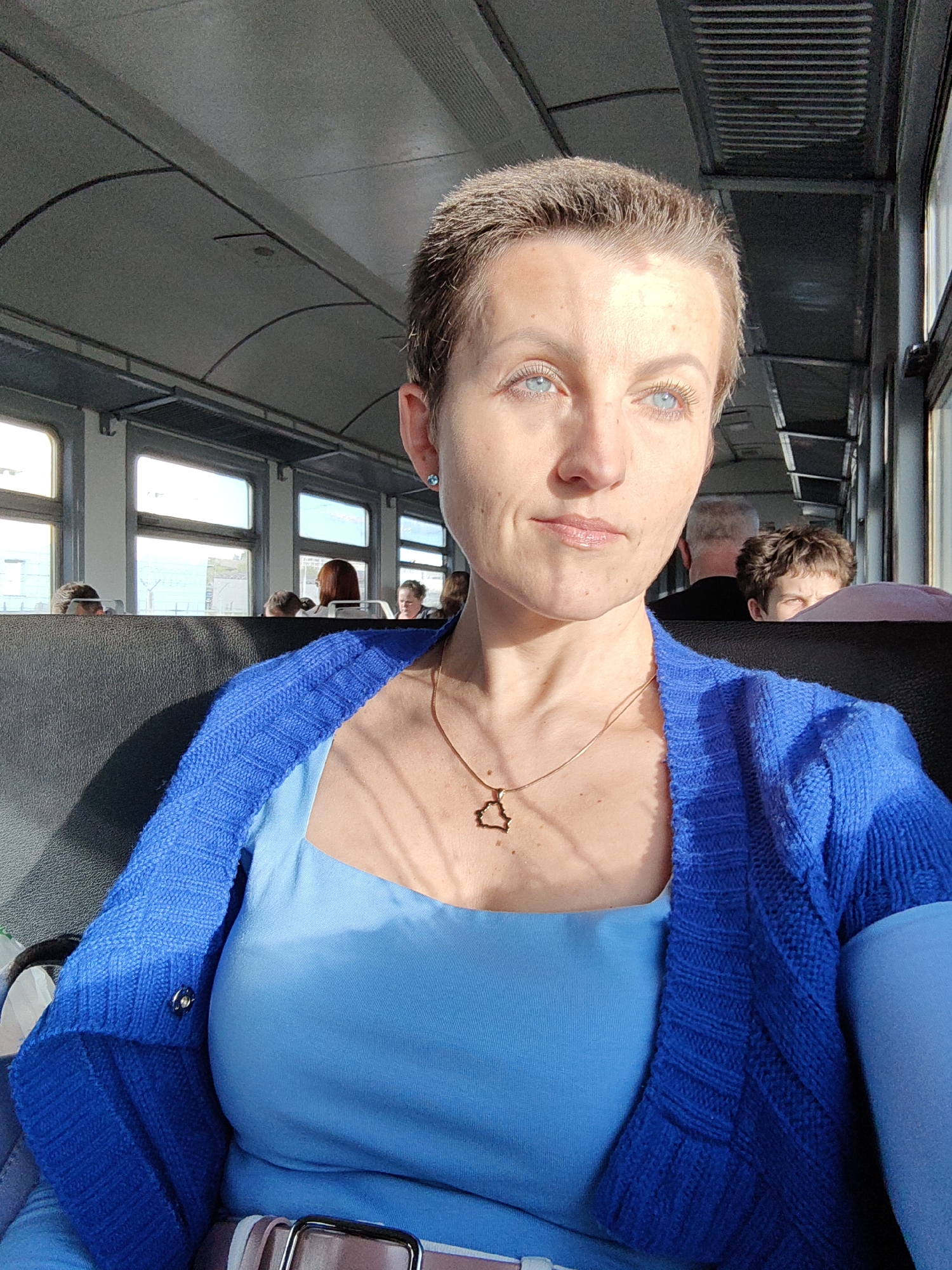 The image shows a close-up, waist-up view of a woman with short, brown hair and blue eyes, looking towards the right side of the frame. She is wearing a bright blue cardigan over a lighter blue top and a thin gold necklace with a geometric pendant. The background appears to be the interior of a train carriage, with windows on the left showing an outdoor scene and other passengers visible in the distance. The lighting suggests it is daytime, with sunlight streaming in from the windows. The overall impression is a candid portrait taken during travel. This scene does not provide any specific details to identify the location as Minsk, Belarus.