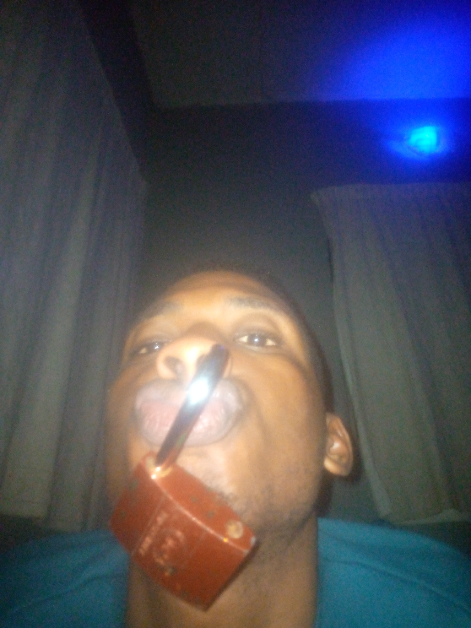A low-angle, dimly lit selfie shows a dark-skinned man with short, close-cropped hair holding a brown, rectangular pipe in his mouth. The stem of the pipe extends upwards towards his nose. His lips are pursed around the pipe. The man is wearing a blue shirt. Behind him are partially drawn white curtains. At the top of the image, there is a blue light source. The location of this image is in the city of Unknown, Unknown.