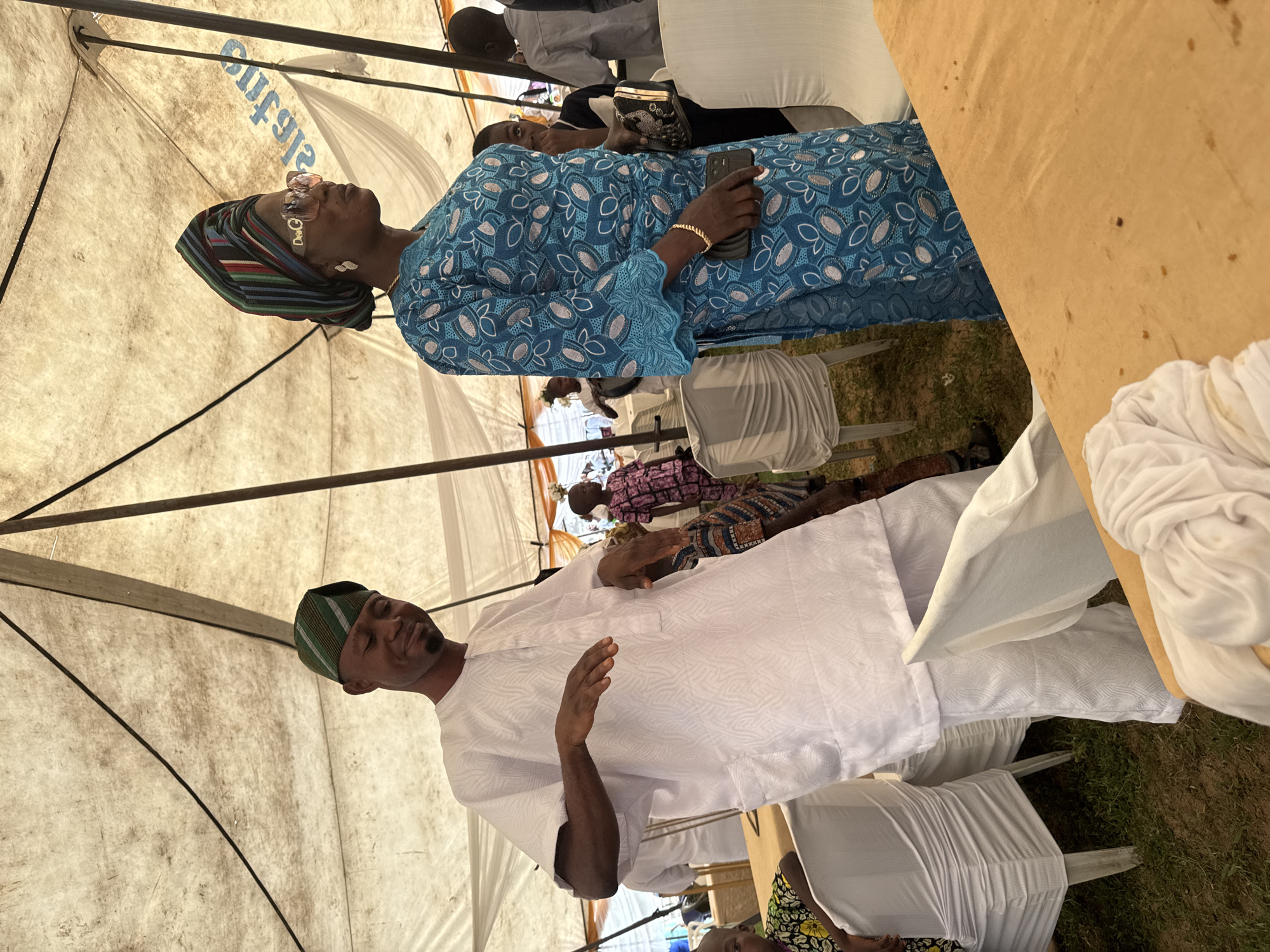 The image depicts an outdoor event under a large, light-colored tent, likely in the city of Badagry, Nigeria. A man in a white traditional outfit and a patterned green cap is gesturing with his hands. Beside him, a woman wears a blue traditional outfit with a patterned design and a colorful headwrap, accessorized with large sunglasses and jewelry. In the background, several white chairs with fabric coverings are visible, along with other individuals, including children. The ground appears to be grass, suggesting a park or open space. The tent's fabric has some writing on it, but it is partially obscured.