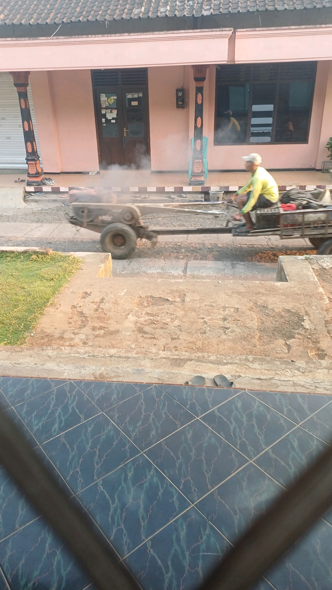 A person wearing a yellow shirt and a white cap is operating a two-wheeled trailer that is being pulled by a small engine. The trailer is loaded with dark material, and steam is rising from it. The scene appears to be a street in Wonokerso, Indonesia, in front of a light pink building with a brown tiled roof. The building has dark brown double doors and windows with reflective glass. The ground in front of the building is paved with concrete, and there is a grassy area to the left. There are also several pairs of flip-flops scattered on the ground near the building. The foreground of the image shows a tiled floor.