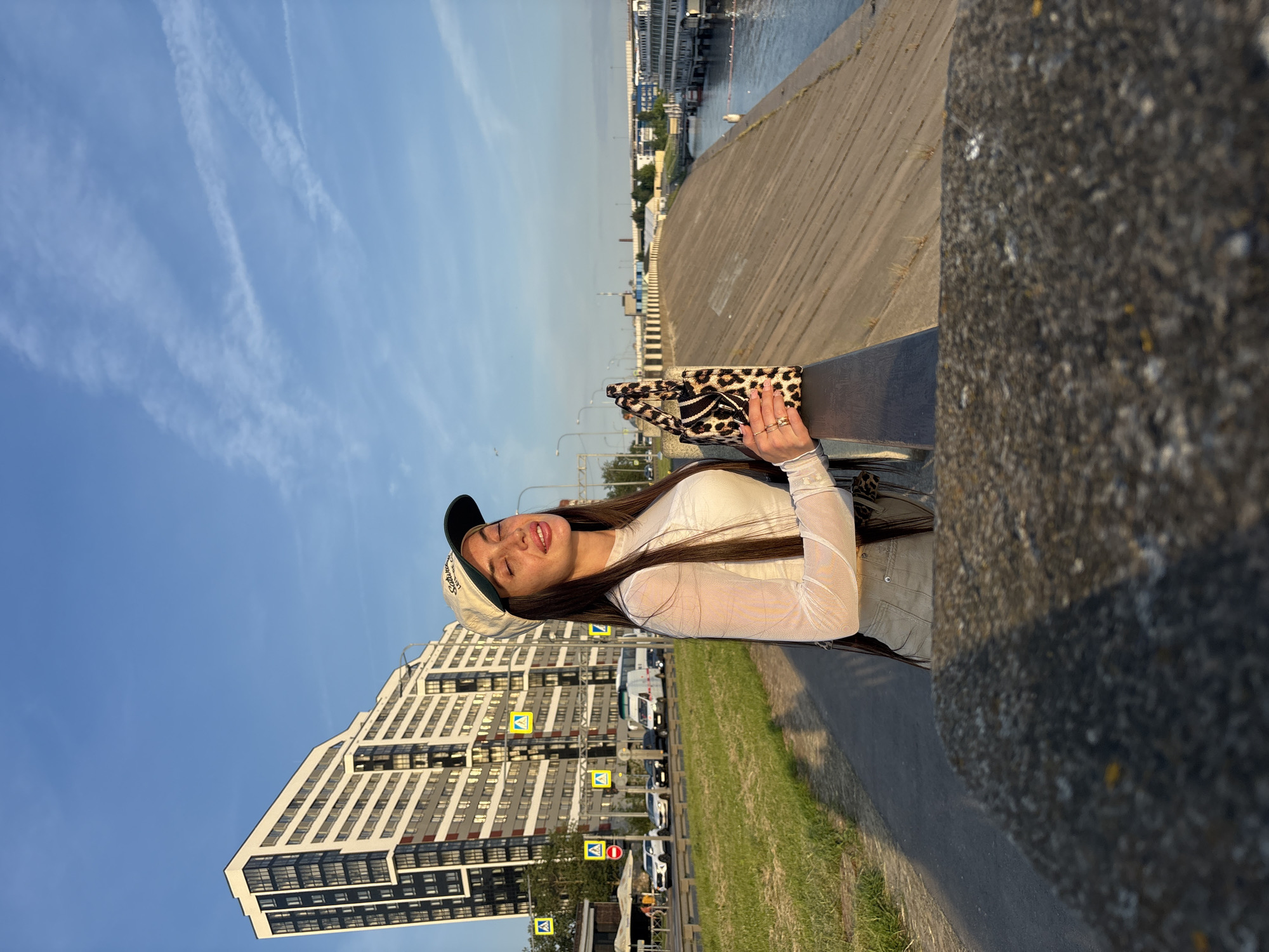 A young woman with long dark hair, wearing a white sheer long-sleeved top and light-colored pants, stands outdoors with her eyes closed and head tilted back, enjoying the sunlight. She is wearing a baseball cap and holding a small leopard-print handbag. The woman is standing on a paved walkway next to a concrete embankment that slopes down to a river. In the background, a modern, multi-story building with many windows is visible on the left, along with a road with several cars and traffic signs. The sky is blue with thin white clouds. The scene appears to be located in Saint Petersburg, Russia, possibly near the Neva River embankment.