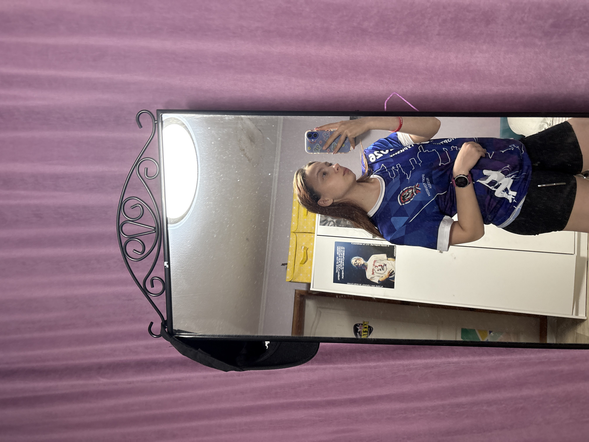 The image shows a woman taking a picture of herself in a mirror. She is wearing a blue soccer jersey, black shorts, and a smartwatch. The mirror is framed in black metal with decorative flourishes, and a black baseball cap hangs from the bottom. The background appears to be a room with a pink wall, a white cabinet with a poster on it. Based on the shirt, the location could be Прикамский, Russia.