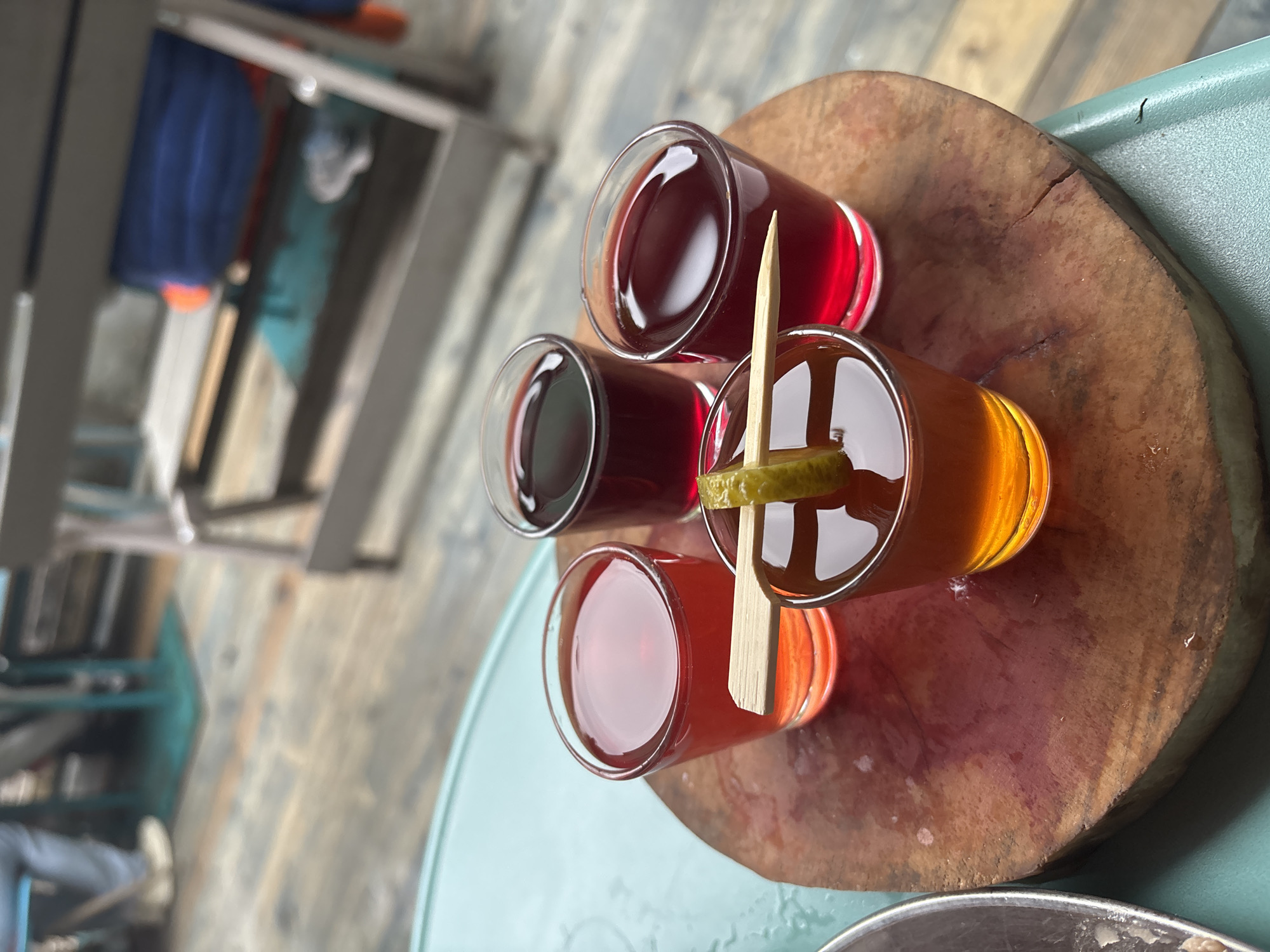 The image shows a wooden cutting board with four small shot glasses of various colored liquids. A small piece of green garnish, possibly a lime or cucumber slice, is speared by a wooden pick and placed across the front shot glass. The shot glasses are arranged in a circular pattern on the board. The background appears to be an outdoor setting with wooden planks on the ground and blurred furniture. No specific location in Komarovo, Russia is identifiable.