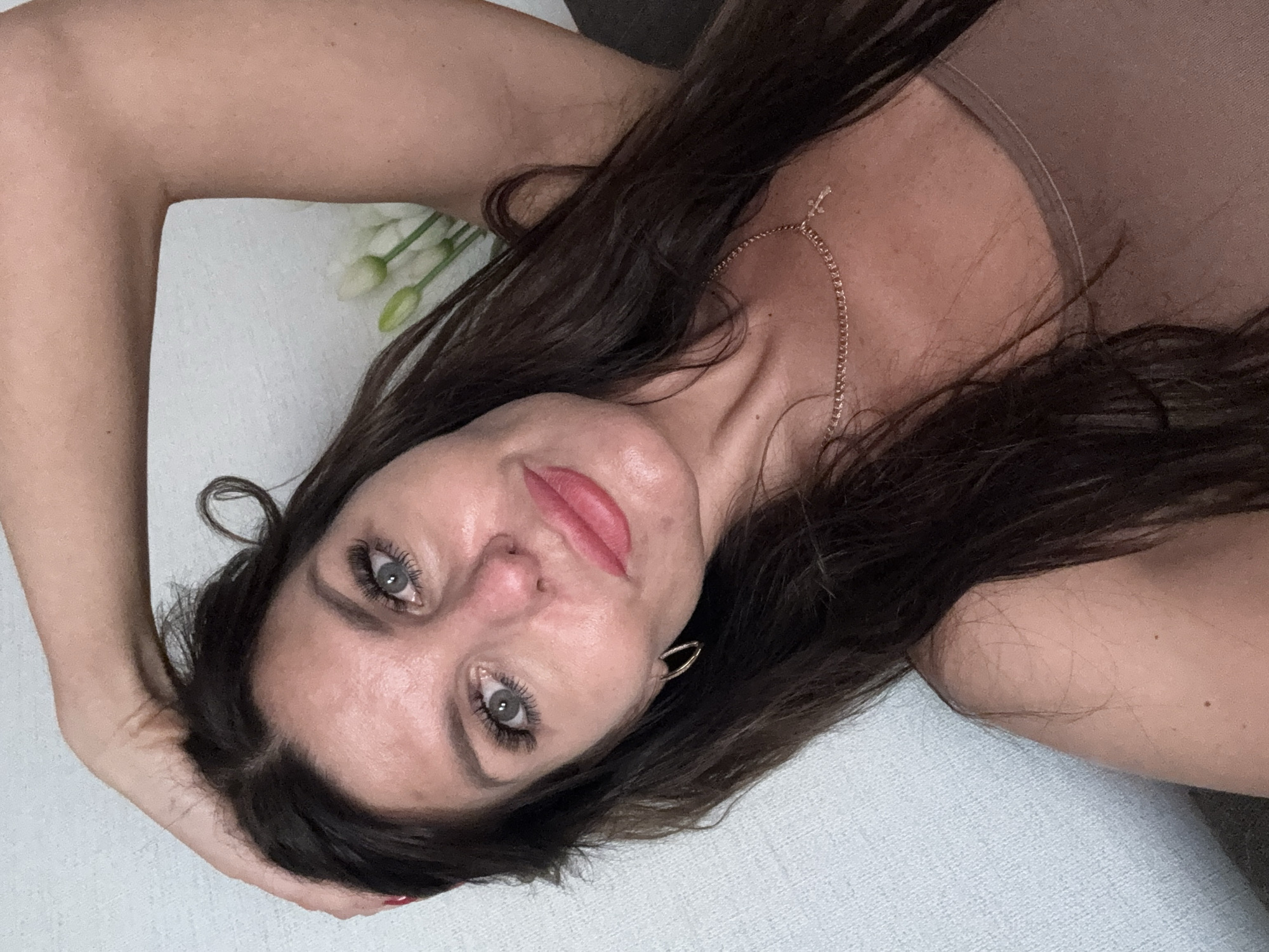 The image shows a medium shot of a woman posing for a selfie. She is wearing a light brown tank top and a gold necklace with a cross pendant. Her right arm is raised and resting on the back of her head, and her left arm is out of frame. She has long, dark brown hair, blue eyes, and is wearing makeup. In the background, there is a light blue textured wall and a blurred bouquet of white flowers. The scene is indoors, likely in a residential setting. The image does not provide any information about the city of Chaikovsky, Russia.