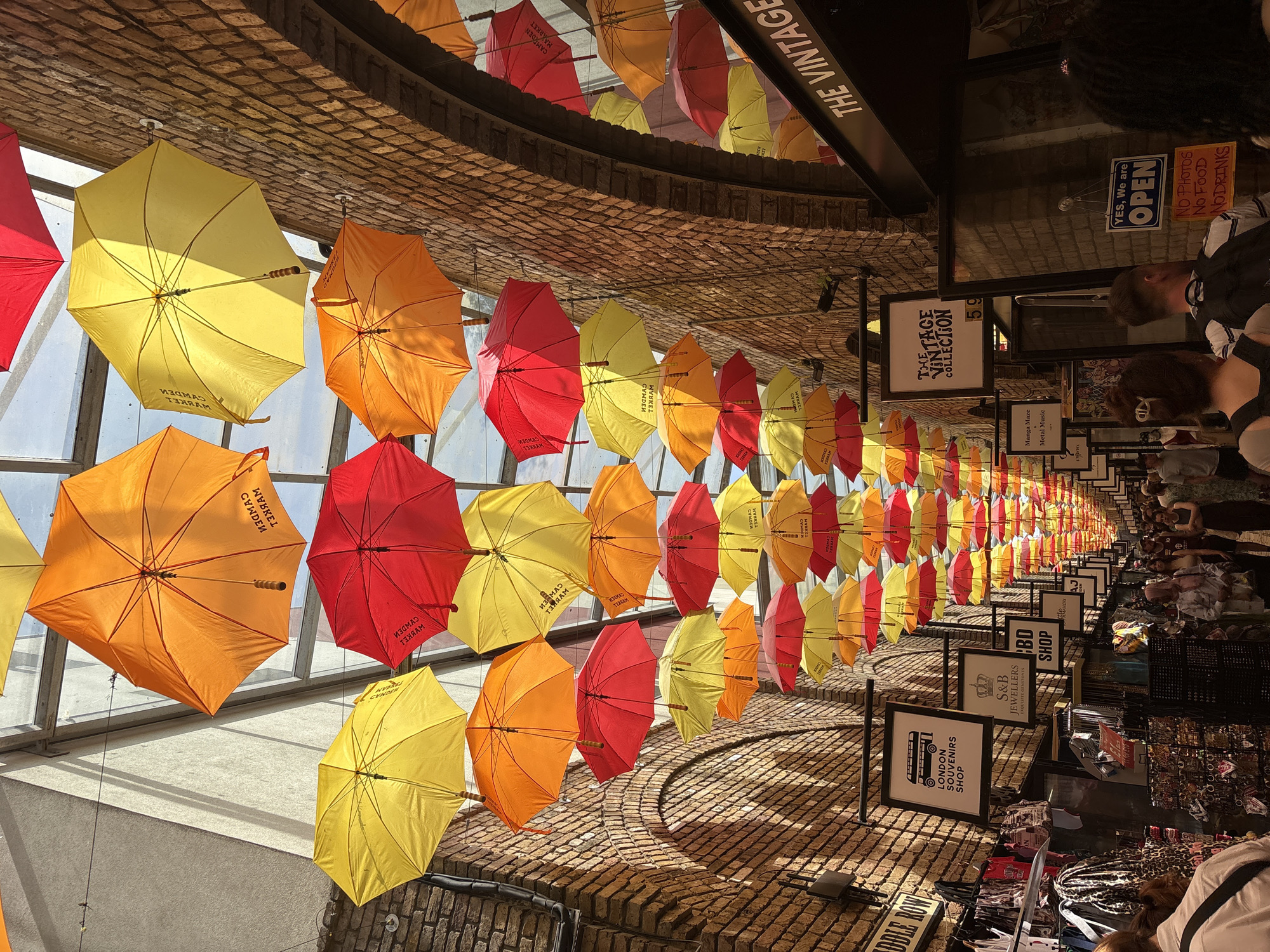 The image shows the interior of Camden Market, in London, United Kingdom. The perspective is looking down a central corridor with vendor stalls lining the sides. Suspended above are rows of colorful, open umbrellas (red, yellow, and orange). Signs hang above the stalls advertising different shops, like "London Souvenirs Shop," "CBD Shop," and "S&B Jewellers." The architecture includes brick archways. Patrons are browsing the merchandise. One sign reads, "The Vintage Collection." Another sign states, "YES, We are OPEN" and "No Photos, No Food and Drinks."