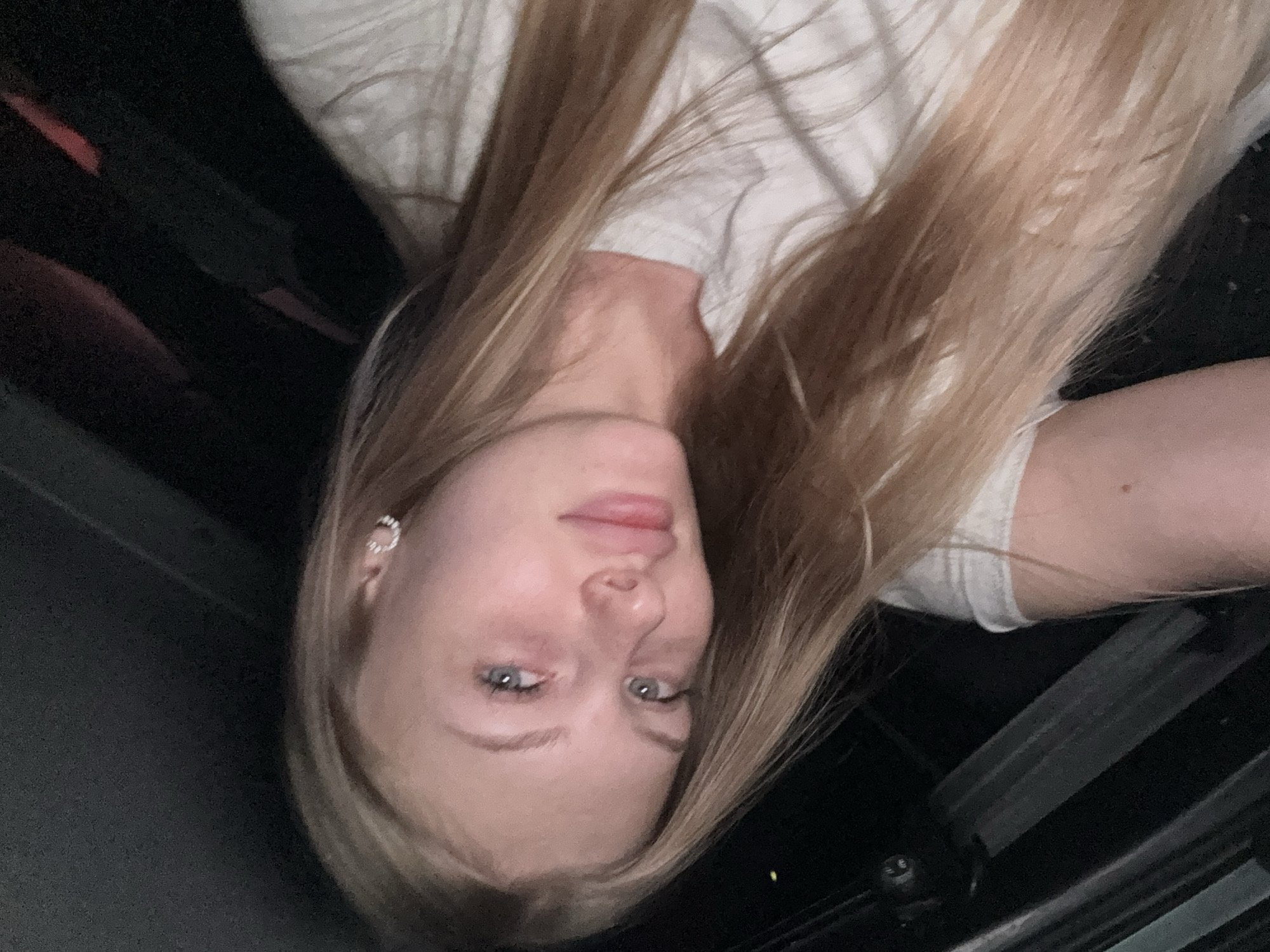 The image shows a close-up, angled selfie of a young woman with long blonde hair. She has blue eyes and is wearing a white t-shirt. A small, circular earring is visible on her left ear. The background is dark and out of focus, suggesting an indoor setting, possibly a car or a dimly lit room. There are no specific landmarks or details that would allow for the identification of the city of Perm, Russia, or any specific location within it.