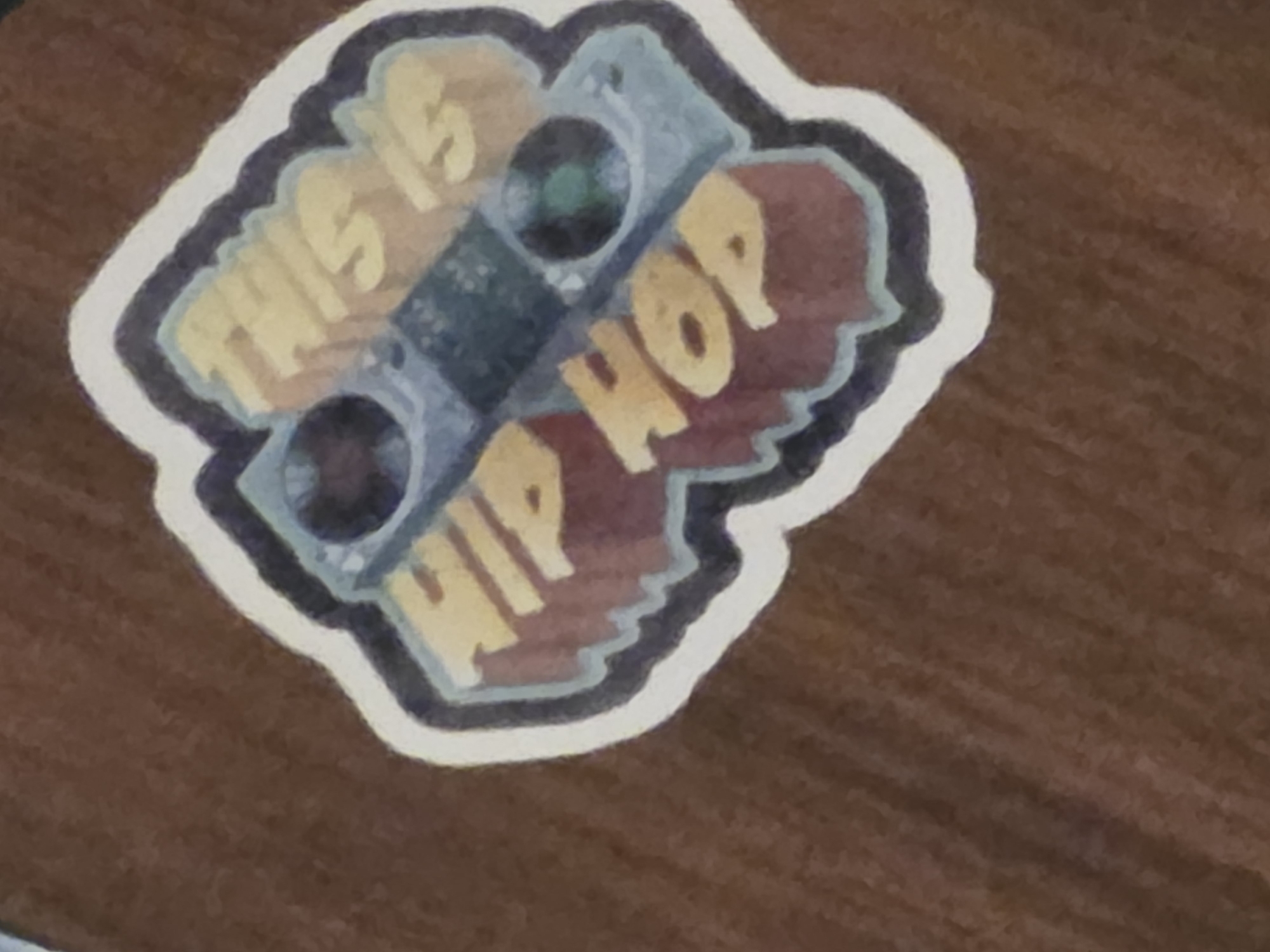 A sticker with the words "THIS IS HIP HOP" is displayed on a brown, textured surface. The sticker features a stylized depiction of DJ equipment, including two turntables and a mixer. The words are presented in a bold, 3D font with yellow lettering and a dark red base. The background of the sticker has a white outline, followed by a dark blue and light blue wavy border. The surface the sticker is on appears to be fabric, possibly clothing, due to its texture and the way it drapes. The lighting suggests an indoor environment, but the exact location within the city of Unknown, Unknown, is not discernible. No people or actions are visible in the image.