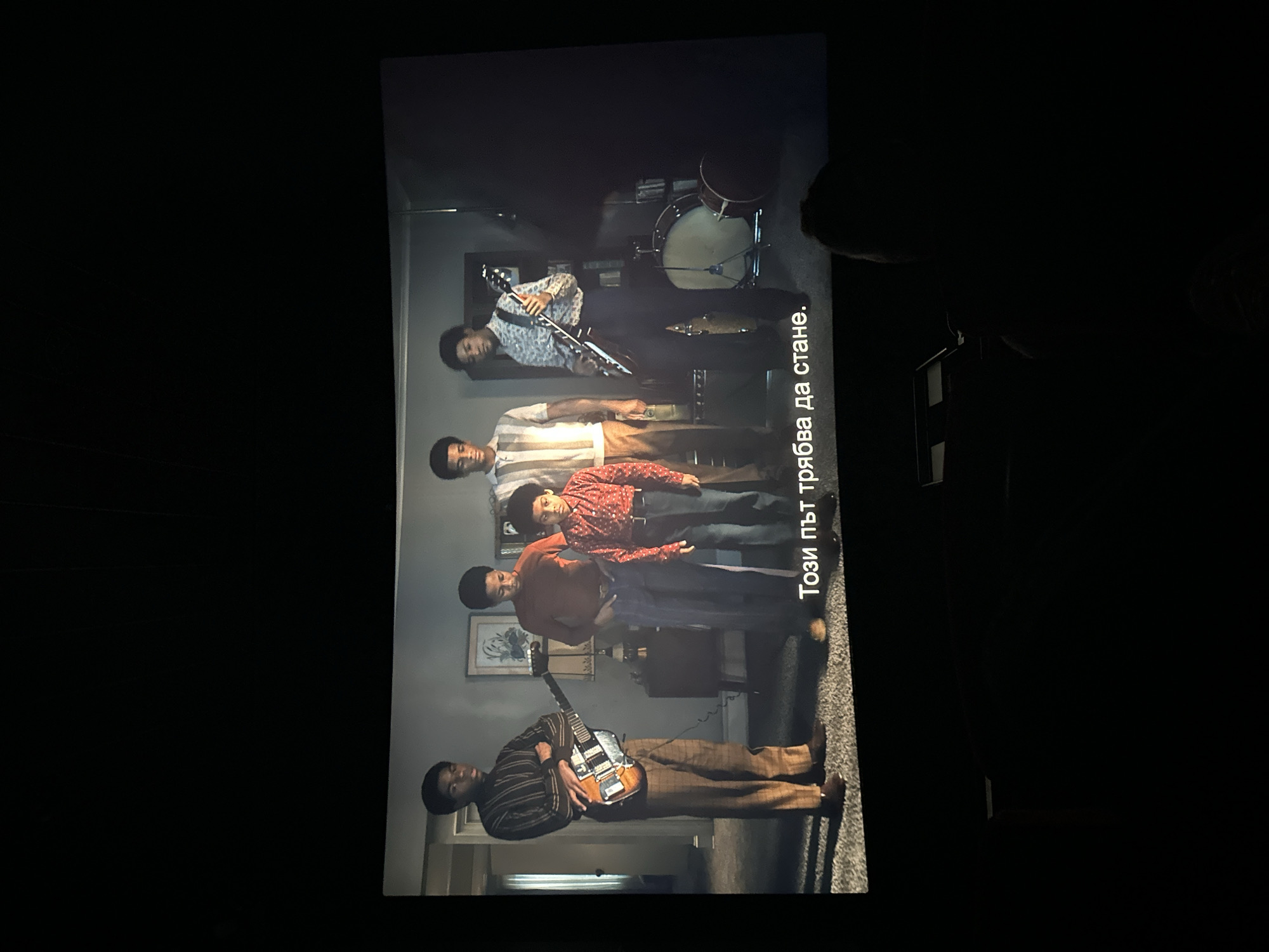 A screen displays a band of five young Black men posing for a photo in a domestic setting. From top to bottom, they are arranged as follows: a drummer behind his kit, a saxophone player, and three others. The lowest person is playing an electric guitar. The background includes a painting on the wall and a glimpse of a doorway. The image has Bulgarian subtitles written on it. The setting does not appear to be in Varna, Bulgaria, but instead in a North American home.
