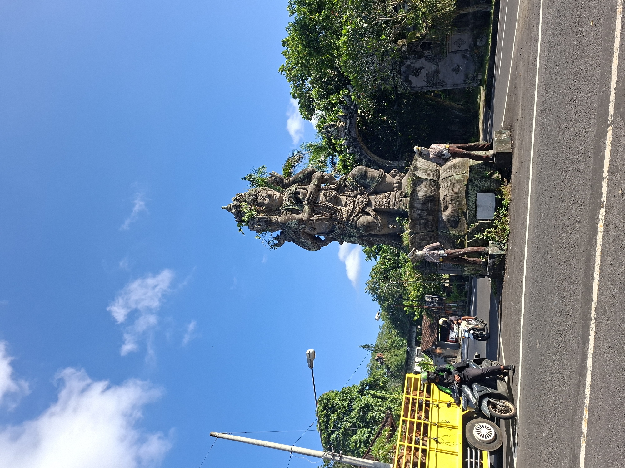In Banjarangkan, Indonesia, a street scene unfolds under a bright blue sky dotted with white clouds. Dominating the center of the frame is a large, weathered stone statue, likely depicting a deity, adorned with multiple arms and intricate headdresses. The statue sits on a tiered stone base, with patches of moss and vines growing on it. Two smaller stone figures, one resembling a police officer in uniform and the other appearing to be a woman, stand at the base of the statue, one on each side.
To the left, a bright yellow truck filled with cattle is stopped at the roadside. A person wearing a green helmet and dark clothing rides a motorcycle in front of the truck, with another person on a white motorcycle behind them. A lamppost stands tall to the left of the truck, and another lamppost is visible further down the road. Lush green trees line the background, with hints of buildings and structures behind them. The road in the foreground is paved and marked with white lines. The overall atmosphere is that of a bustling street in a tropical location.