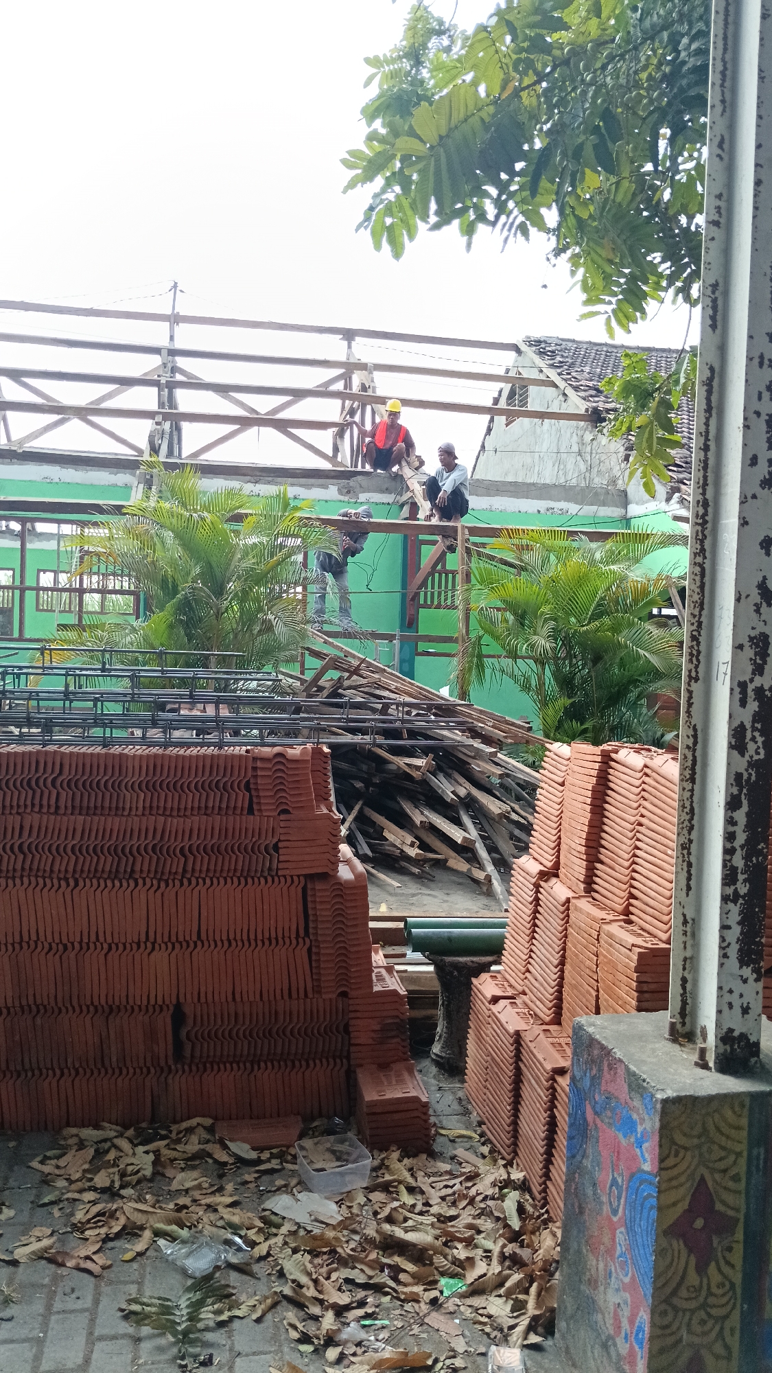 The image captures an outdoor scene at a construction site in Branggahan, Indonesia, likely a residential or school building undergoing renovation or repair. Three male construction workers are present. One worker, wearing a yellow hard hat and an orange vest, is seated on wooden beams near the roofline. Another worker, dressed in a grey shirt and dark pants, is also seated on the beams, facing the first worker. A third worker, wearing a grey long-sleeved shirt and dark pants, is standing on a higher wooden platform and appears to be engaged in work, possibly using tools.
The foreground of the image is dominated by stacks of terracotta roof tiles, arranged neatly in multiple rows. Adjacent to the tiles, a pile of wooden planks and construction debris is visible. Several palm trees and other green foliage are interspersed throughout the scene, suggesting a tropical or subtropical environment. In the background, a building with a green facade and visible windows can be seen. The sky is overcast. The overall impression is one of ongoing construction work under a cloudy sky.