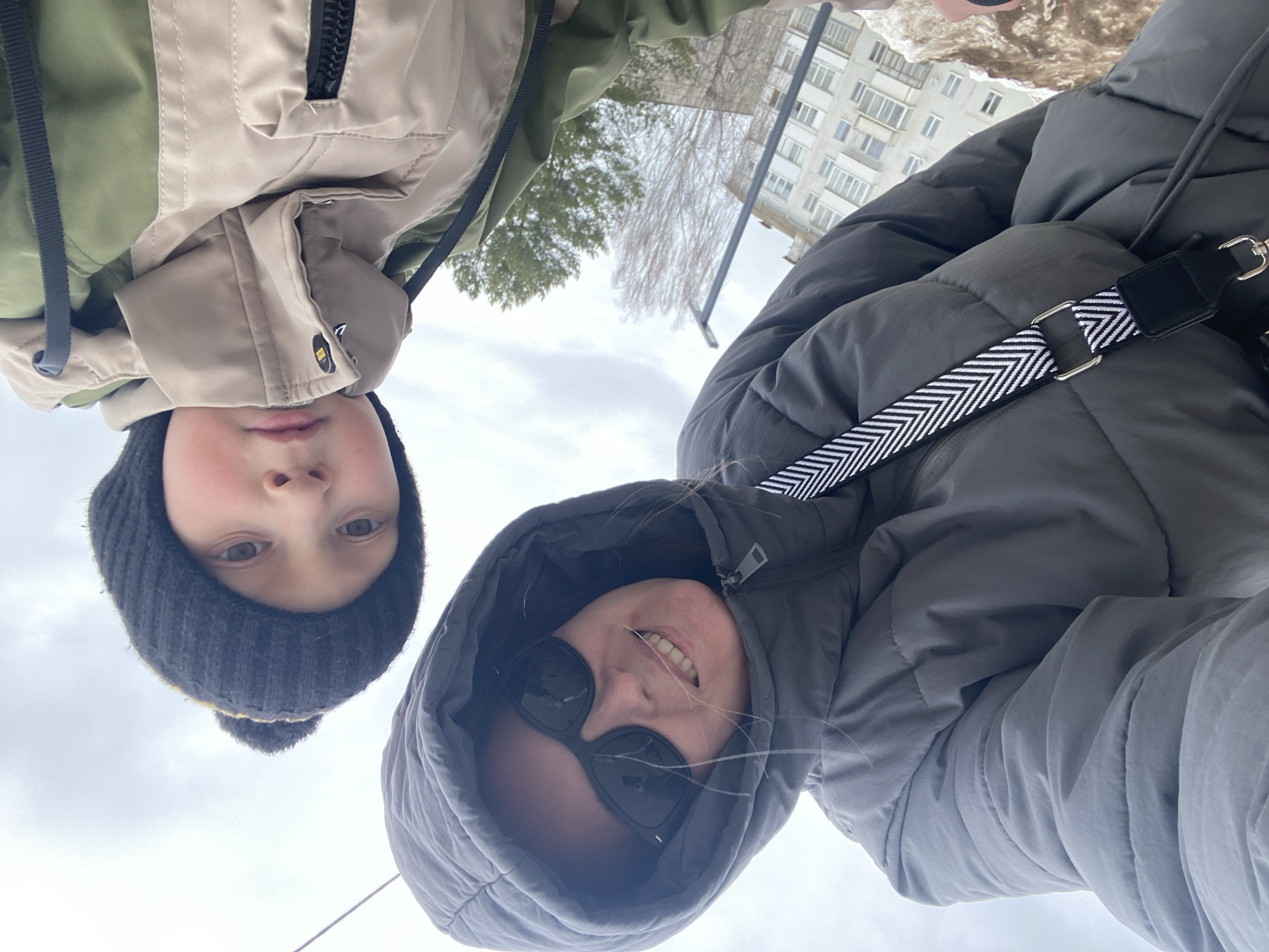 Two people are shown from an unusual perspective, as if they are looking down at the viewer. A child in a dark knit hat with a yellow stripe is on the left. To the right is an adult wearing sunglasses and a hooded gray puffer jacket, a camera strap visible across the chest. The background includes a multi-story apartment building and a cloudy sky. The building is located in Noviy, Russia.