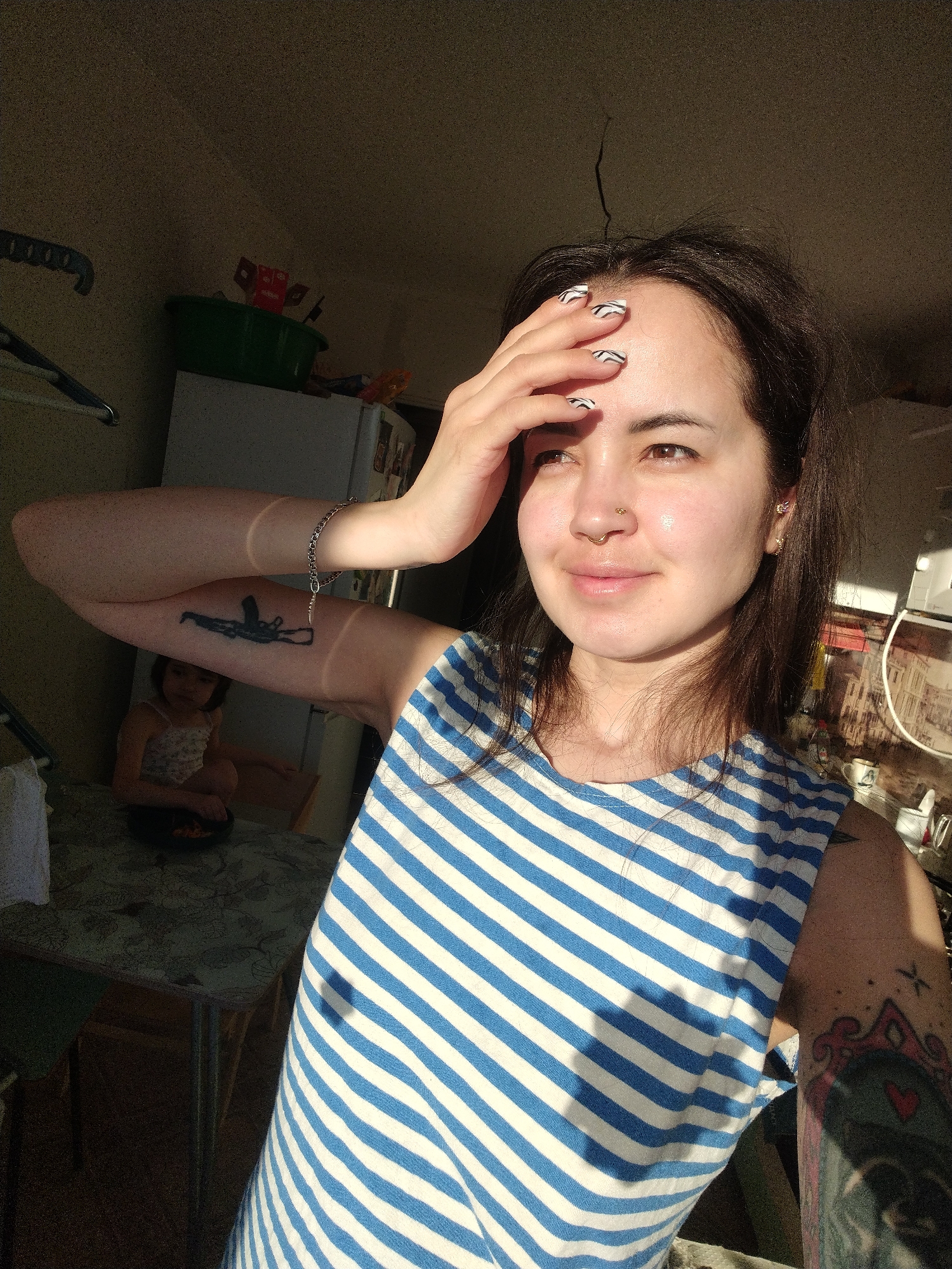 This indoor photograph, taken in Noviy, Russia, features a woman in the foreground shielding her eyes from bright sunlight, with a young child visible in the background.
The woman, positioned centrally, has medium-length dark hair, fair skin, and brown eyes. She wears a blue and white striped sleeveless top. Her left hand is raised to her forehead, fingers spread, casting a shadow across her brow. Her fingernails have a distinctive black and white zebra-like pattern. She has a gold septum piercing and multiple ear piercings, including a gold stud and an ear cuff. Visible tattoos include a black outline of an AK-47 assault rifle on her left bicep and a colorful, intricate design with a red heart on her right bicep. She has a slight, soft smile, gazing upwards and to the right, possibly towards the light source.
In the blurred background, a young child with dark hair is seated at a table with a green patterned tablecloth, holding a dark bowl. Behind the child, a white refrigerator is visible, topped with a green plastic tub containing several small red and white packets. On the far left, a blue clothes hanger hangs from a hook. The room appears to be a domestic setting, possibly a kitchen or dining area.
The strong, warm sunlight streams in from the right, illuminating the woman's face, hair, and parts of her arms, creating stark shadows across her body and the room. The time of day is clearly daytime, given the intense natural light. There is no visible text in the image.