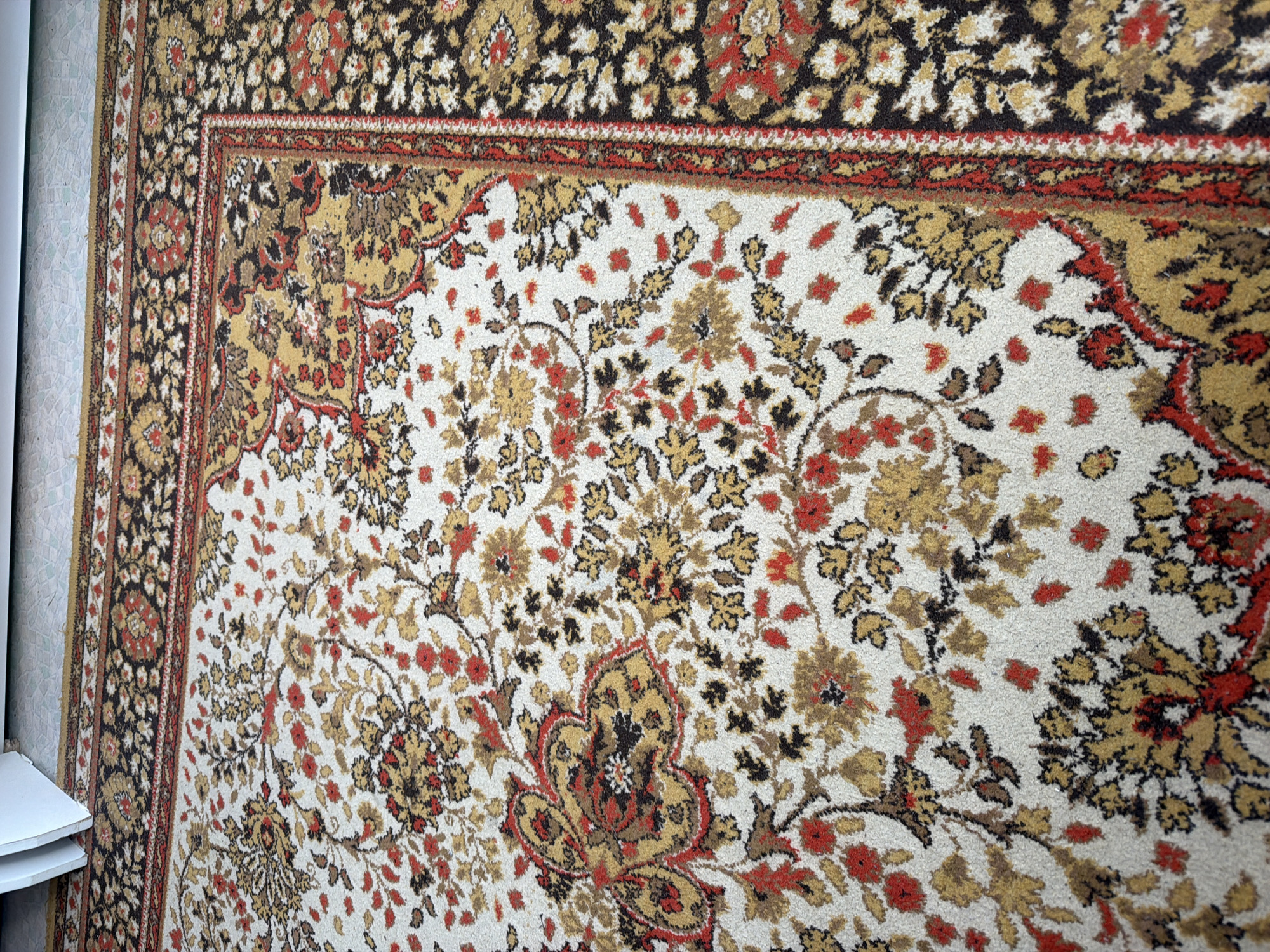 The image displays a close-up view of a patterned rug or carpet, likely hanging vertically on a wall. The rug is adorned with an intricate, ornate floral design.
The dominant background color of the rug's central field is off-white or cream. This lighter background is covered with scattered floral sprays and larger stylized motifs in shades of gold, mustard yellow, reddish-brown (rust), and dark brown or black. The flowers and leaves vary in size and detail, creating a busy and traditional aesthetic. One prominent stylized motif, possibly a paisley or medallion, is visible towards the bottom right of the main field, featuring an elaborate design in gold, red, and dark brown.
The rug has a distinct border that runs along the top and left side of the visible portion. This border is predominantly dark brown or black, also filled with smaller golden and reddish floral patterns. It is framed by narrower stripes in red and gold, separating it from the main field and the very edge of the rug.
On the far left side of the image, a small portion of a white, stepped shelf or furniture piece is visible. Behind this shelf, and continuing along the left edge beyond the rug's border, there is a textured wall surface, possibly wallpaper, in a light, neutral color with a subtle pattern.
The rug has a short, soft pile, giving it a tactile appearance. The lighting appears even, suggesting an indoor setting. There is no visible activity or event taking place, as the image focuses solely on the static rug and its immediate surroundings. No text is visible in the image.