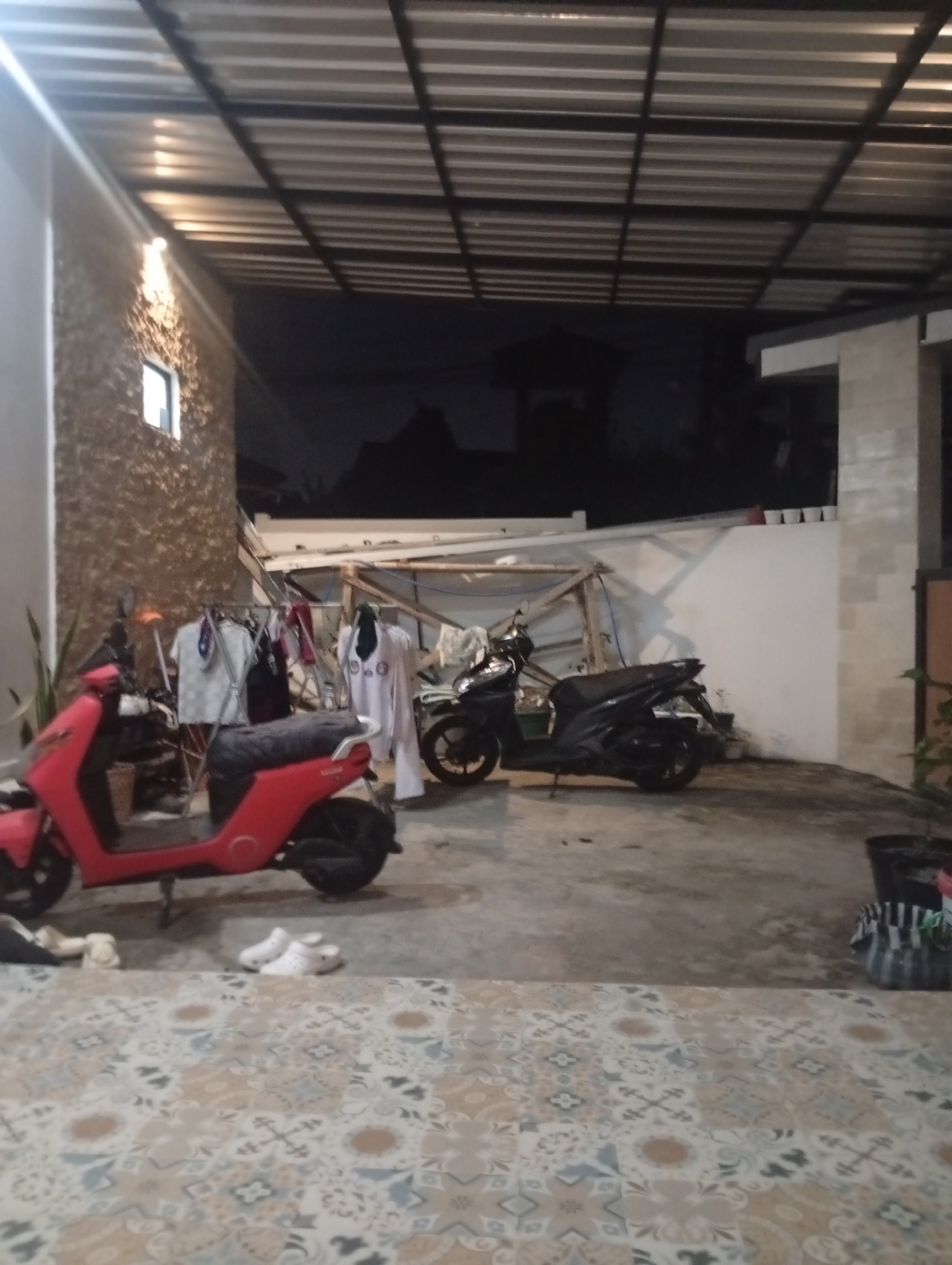 In the city of Denpasar, Indonesia, at night, a covered outdoor area features two parked motorbikes and a clothes drying rack with clothing. The area is illuminated by a single overhead light. The ground is tiled with a patterned design. A red scooter is parked on the left, and a black scooter is parked on the right. The background shows a textured wall and a white wall.