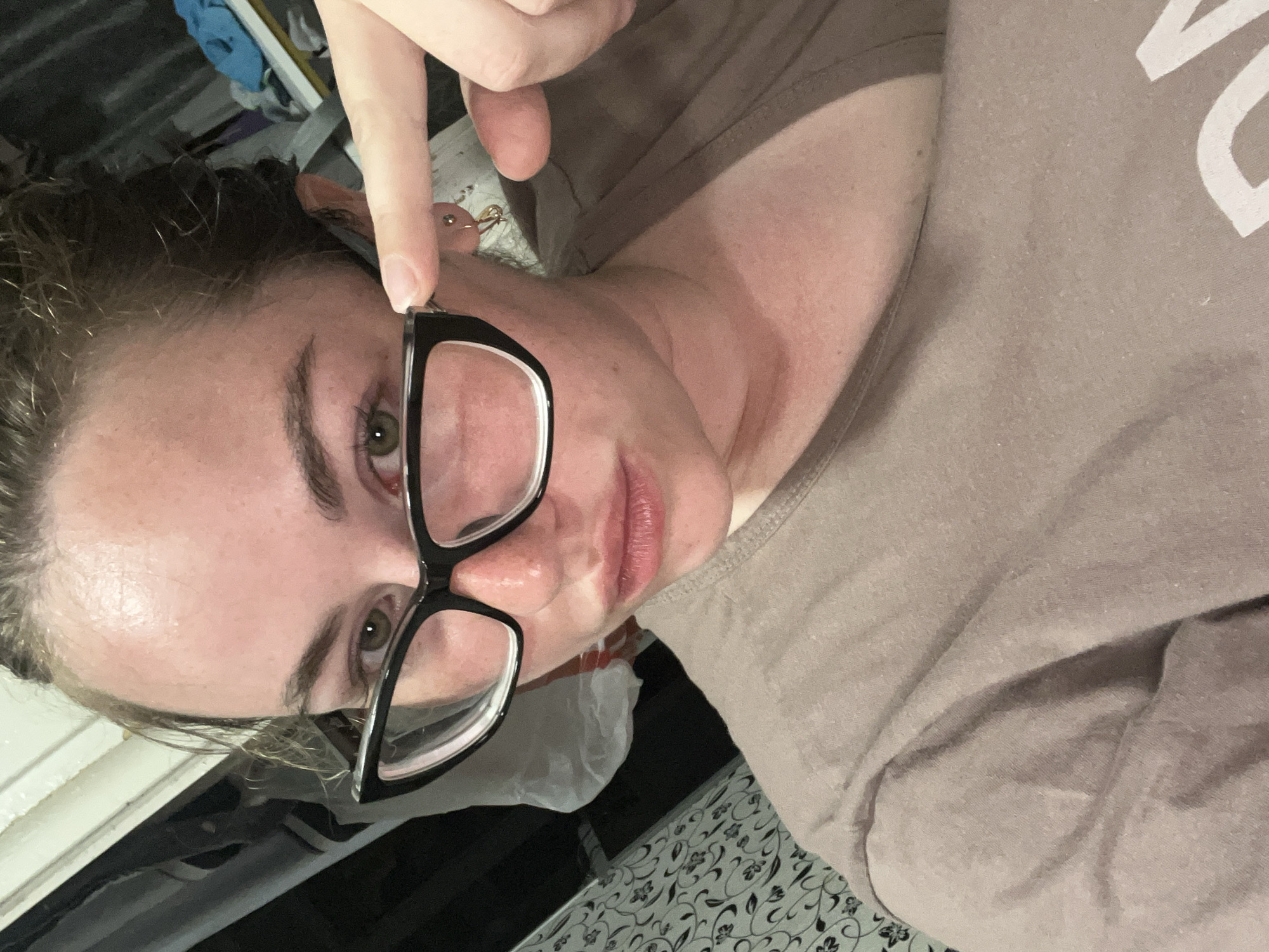 A woman with fair skin and light brown hair is holding a pair of black-framed glasses, pushing them slightly away from her face with her finger. She is wearing a tan-colored t-shirt. The background includes a white plastic bag and a patterned surface. The location cannot be identified from the image.