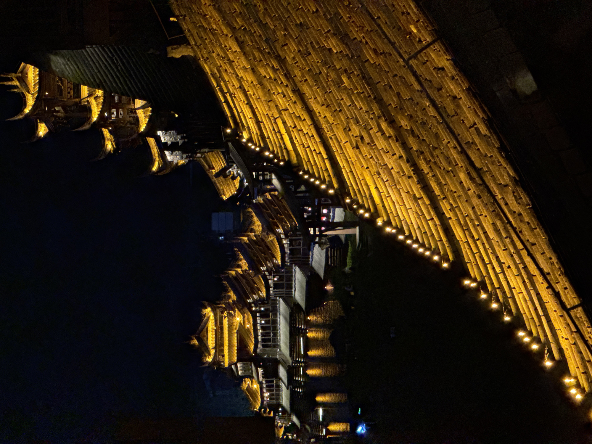 The image shows illuminated buildings at night in Zhuoshui, China. A large bamboo structure with strung lights is positioned at the top right. Multiple buildings are visible along a dark body of water on the left side of the image. The buildings have ornate rooflines. Reflections of the lights are visible in the water.