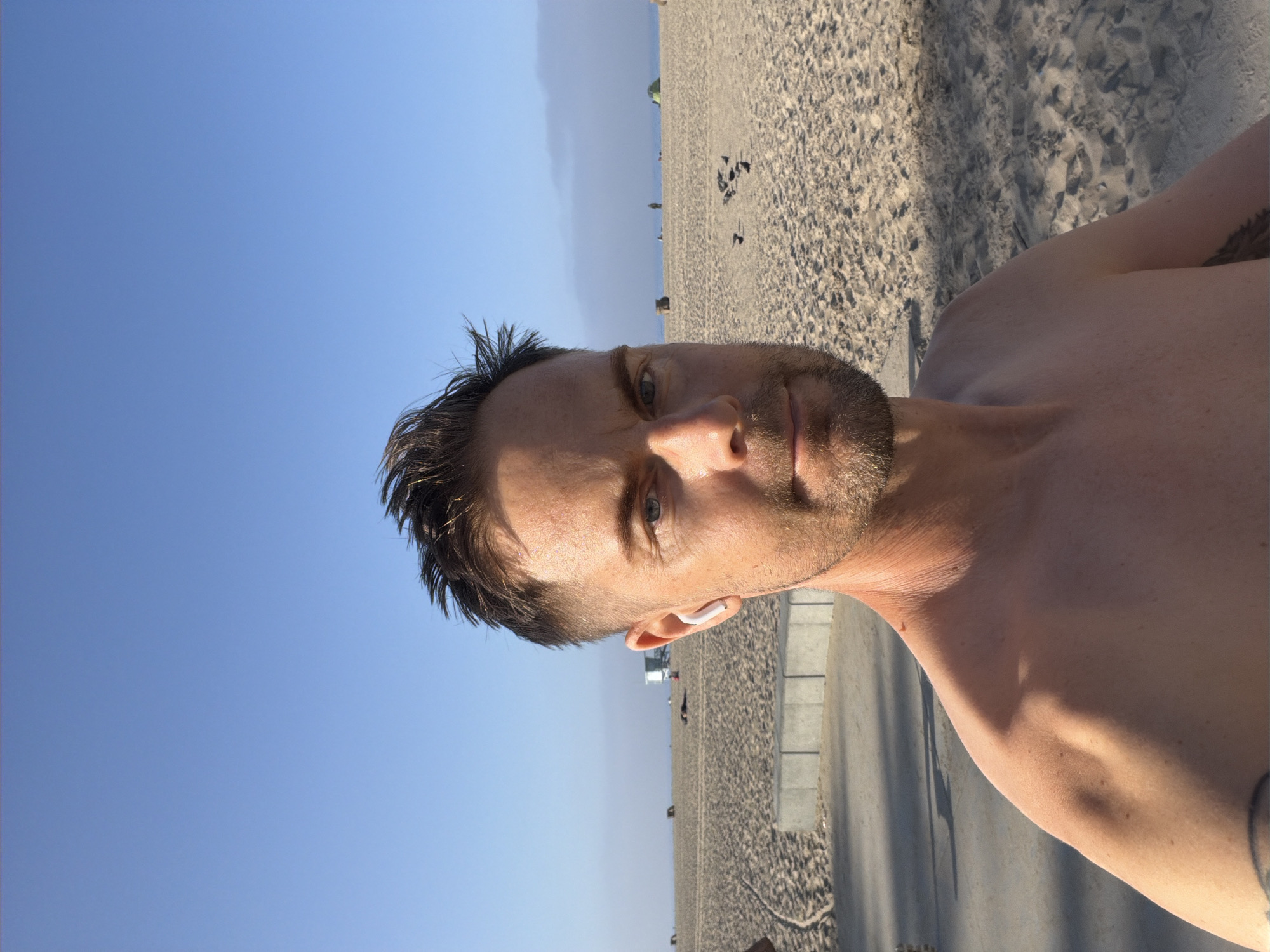 The image shows a man standing on a sandy beach in Santa Monica, United States of America. He is topless and has short, spiked brown hair and a beard. He is wearing white wireless earbuds. The beach is wide and sandy, with some people and tents visible in the distance. The ocean is visible in the background, with a clear blue sky overhead. There is a concrete path behind the man, and some shadows are cast on the sand.