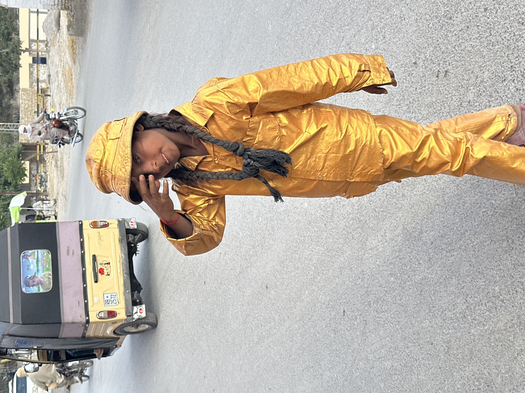 A child in a golden outfit stands on a road in Quetta City Tehsil, Pakistan. The child wears a matching golden hat and has long, dark braids tied together with black string. They look toward the camera, with one hand near their face. In the background, a yellow auto-rickshaw with the license plate "RB 2572" is parked. Further away, a motorcycle with two people on it is visible. The road is asphalt, and buildings are seen in the far background.