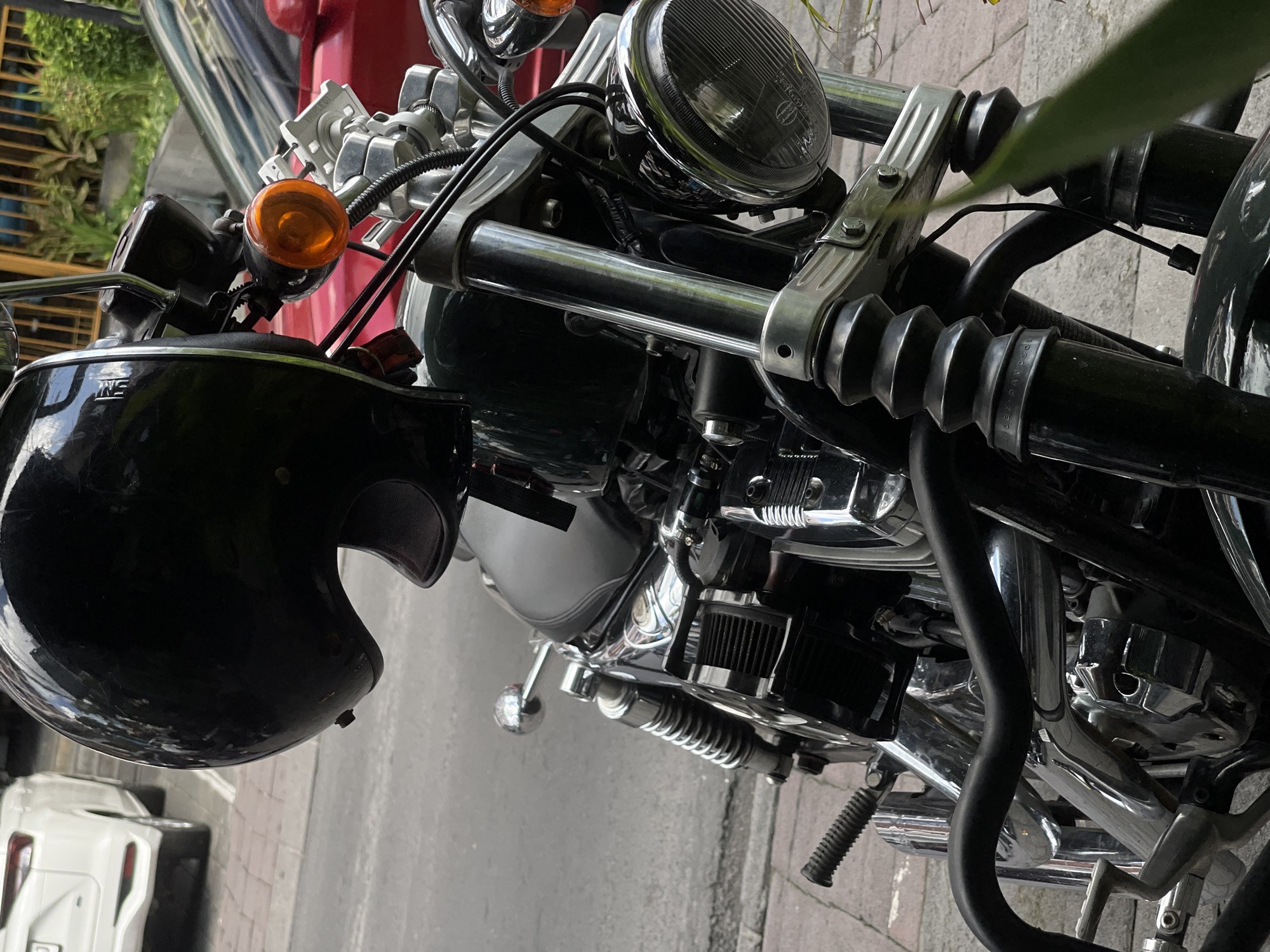 A black motorcycle is parked on a sidewalk in North Kuta, Indonesia. The motorcycle has a black helmet hanging from its handlebars. In the background, a white car is parked on the street. The scene is captured from a medium shot, focusing on the front of the motorcycle. The sidewalk is paved with interlocking concrete blocks, and a gray asphalt street is visible beyond. Some greenery can be seen in the background.