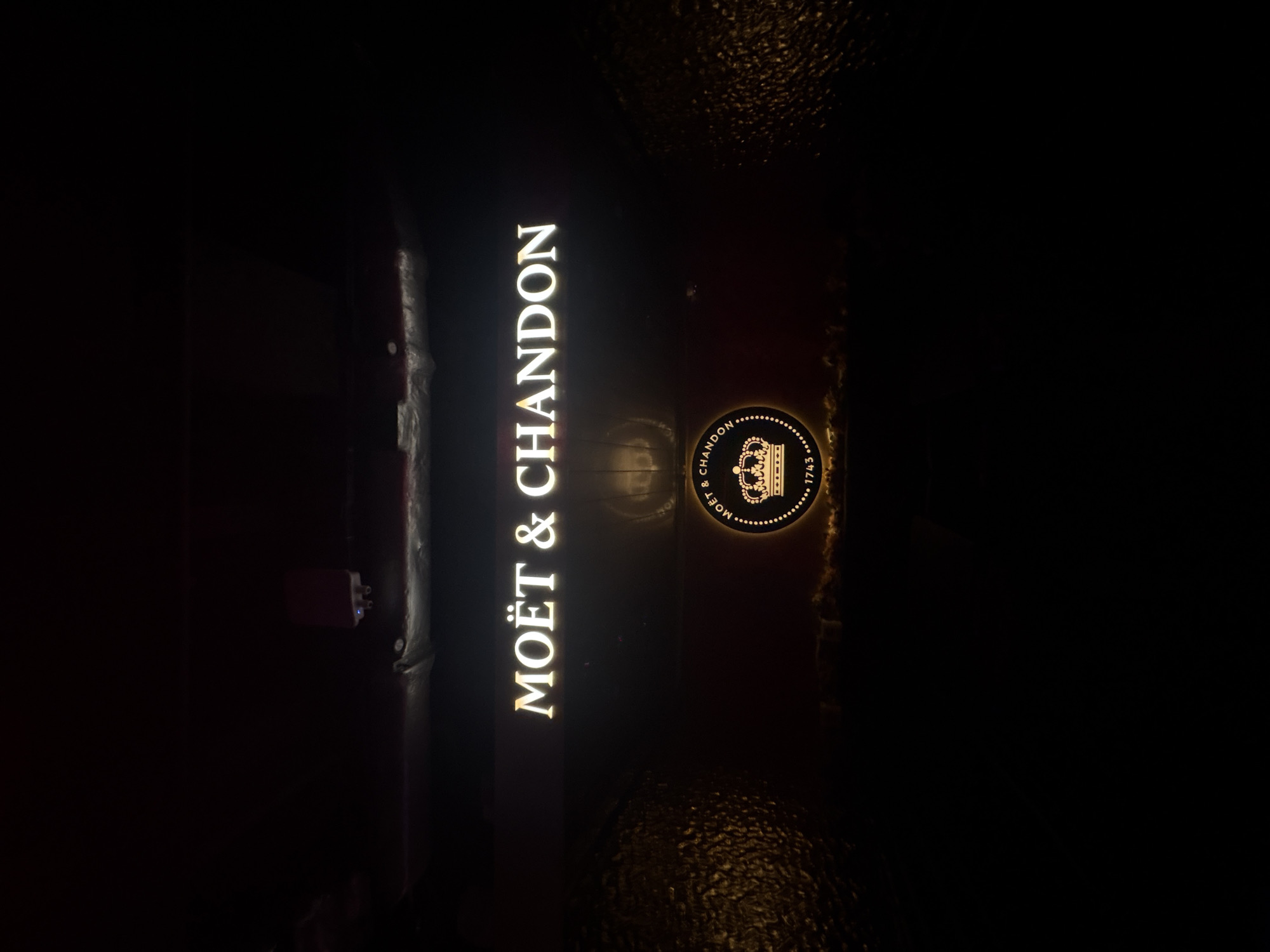 The image shows a vertically oriented, illuminated sign for "MOËT & CHANDON". The sign is attached to a dark surface that could be a wall or facade. The lighting emphasizes the brand name and logo (a crown) in a golden hue. The setting seems to be outdoors at night. The image might have been captured near or in a bar or restaurant in Hong Kong, China, but there is insufficient detail to definitively confirm the specific location within the city.