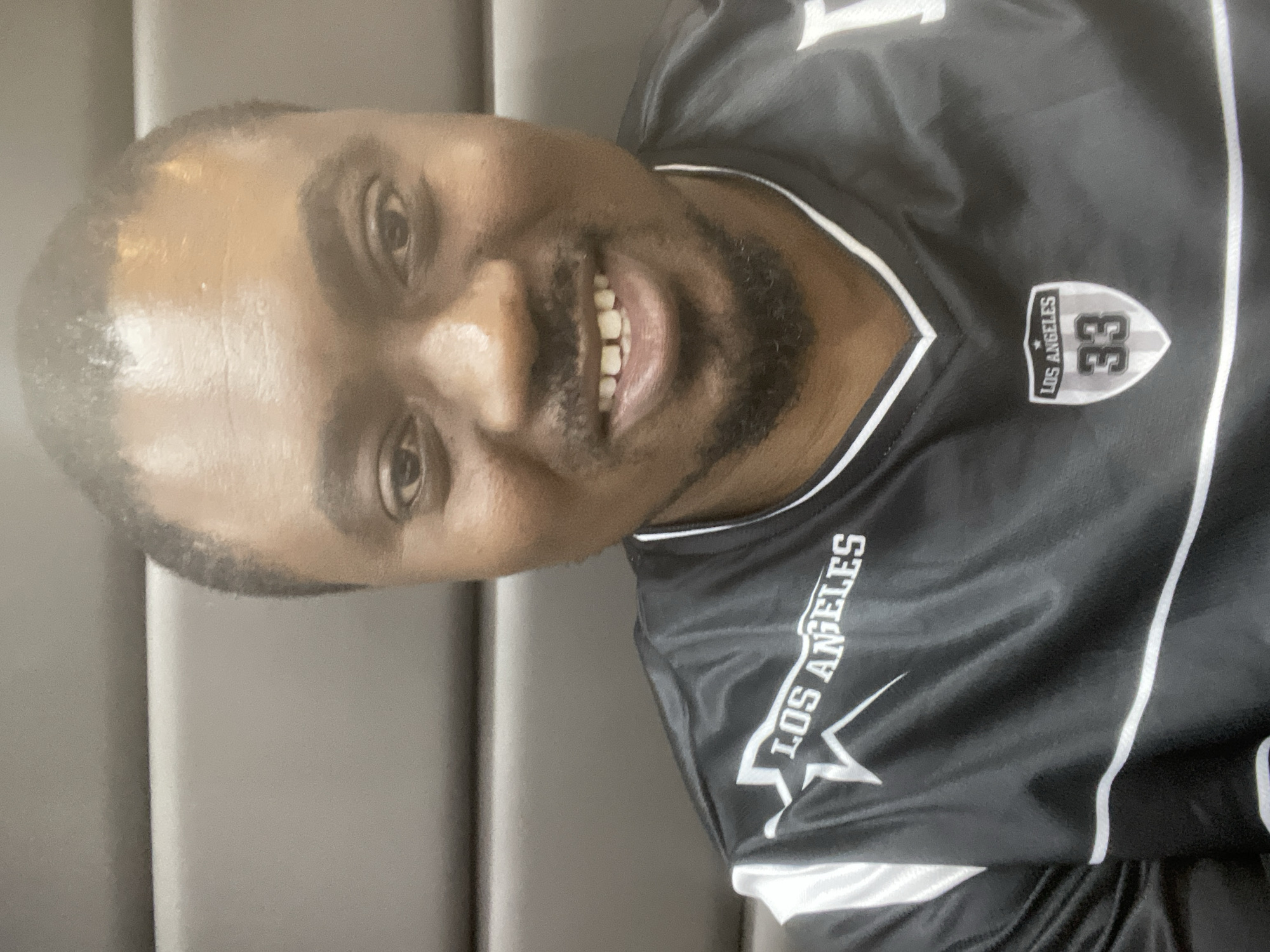 A man with dark skin, wearing a black and white jersey with "Los Angeles" printed on the front and the number 33 on the chest, smiles directly at the camera. He has a trimmed beard and a shaved head with a receding hairline. The background appears to be a gray, possibly upholstered, surface. The image shows an indoor setting and there is no indication of the city of Jos, Nigeria.