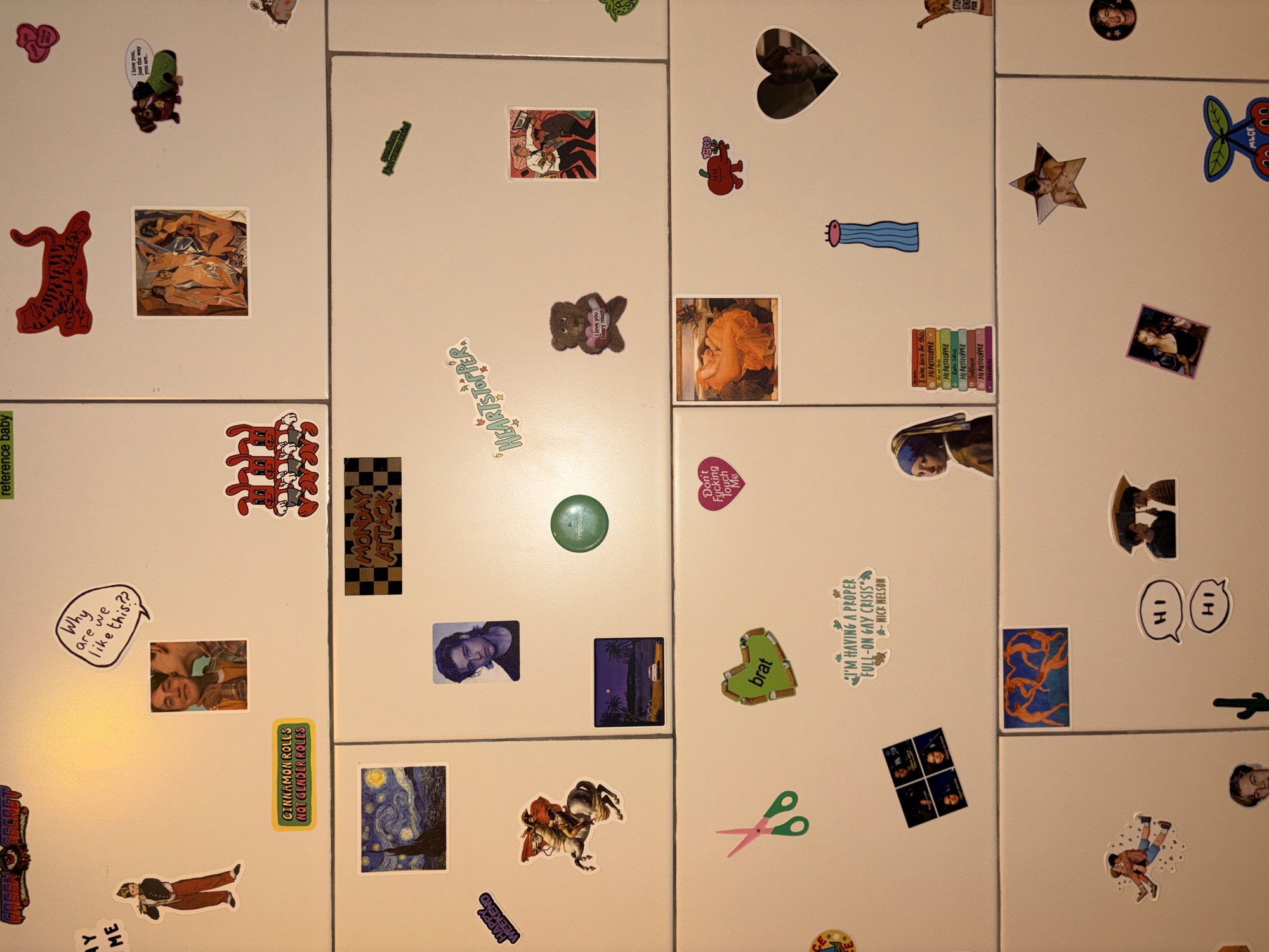 The image shows a close-up, overhead view of a white refrigerator door covered with various stickers. The stickers feature a range of images, including cartoon characters, artwork reproductions, photographs of people, and text-based designs. There are no visible elements that indicate a specific location in Chang Phueak, Thailand. The scene is a static display of decorative stickers on an appliance.