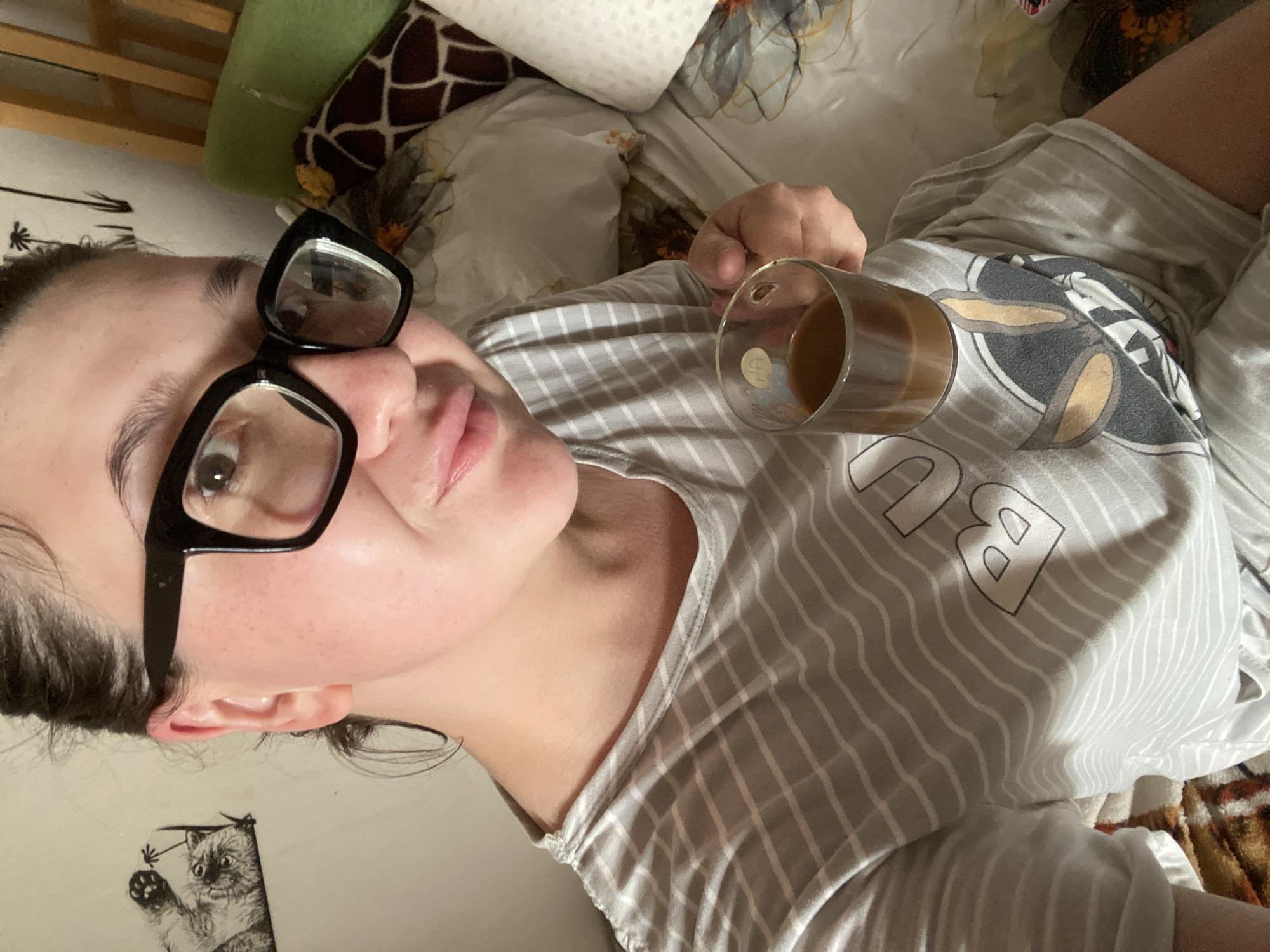 A young woman with dark hair and glasses is holding a glass of coffee while sitting on a bed. She is wearing a striped t-shirt with a cartoon character on it. The room appears to be a bedroom with pillows and blankets on the bed. The background shows a wall with some artwork and a wooden headboard. It is not possible to determine the specific location within Chaikovsky, Russia, from the image alone.