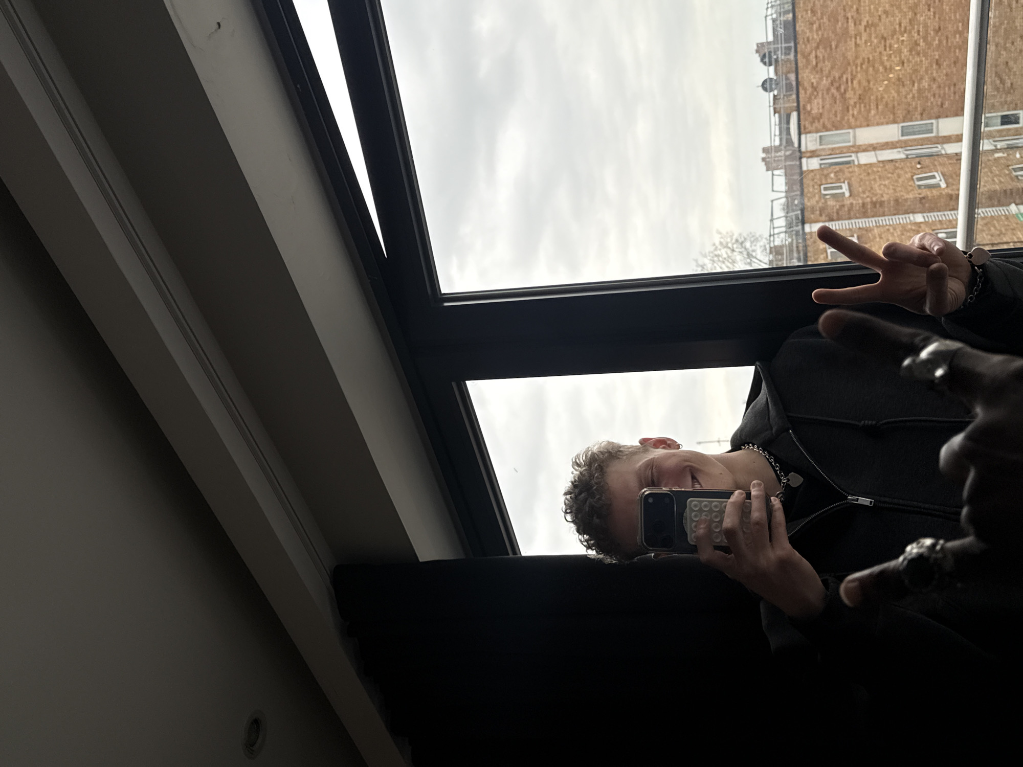 A person is taking a selfie in front of a window, displaying a peace sign with their left hand. They are holding a smartphone in their right hand. The scene is set against the backdrop of a cloudy sky and a building with multiple windows. The location is in the city of London, United Kingdom.