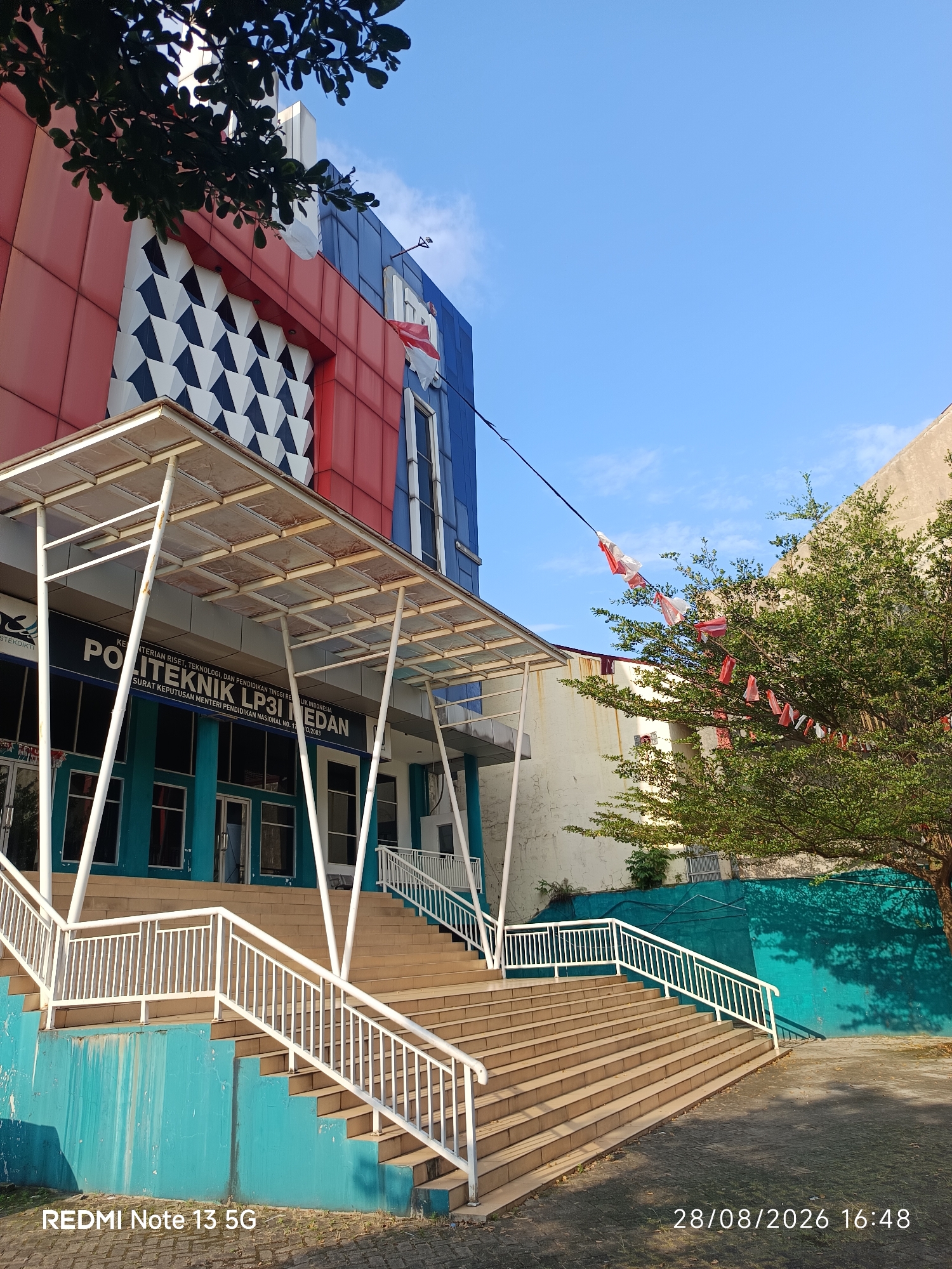 The image shows the exterior of a building in the city of Medan, Indonesia, identified by the text on the building as "POLITEKNIK LP3I MEDAN". The building has a distinctive facade with red and blue sections, and a geometric pattern of black and white shapes. A covered walkway with a metal roof extends over a wide set of outdoor stairs. White railings line the stairs. To the right of the stairs and building, a tree with green foliage is visible, and a string of red and white flags is draped across a wire. The sky is clear blue. The bottom of the image contains text identifying the device used to take the photo as "REDMI Note 13 5G" and the date and time as "28/08/2026 16:48". There are no people visible in the image. The environment appears to be an outdoor area in front of an educational institution.