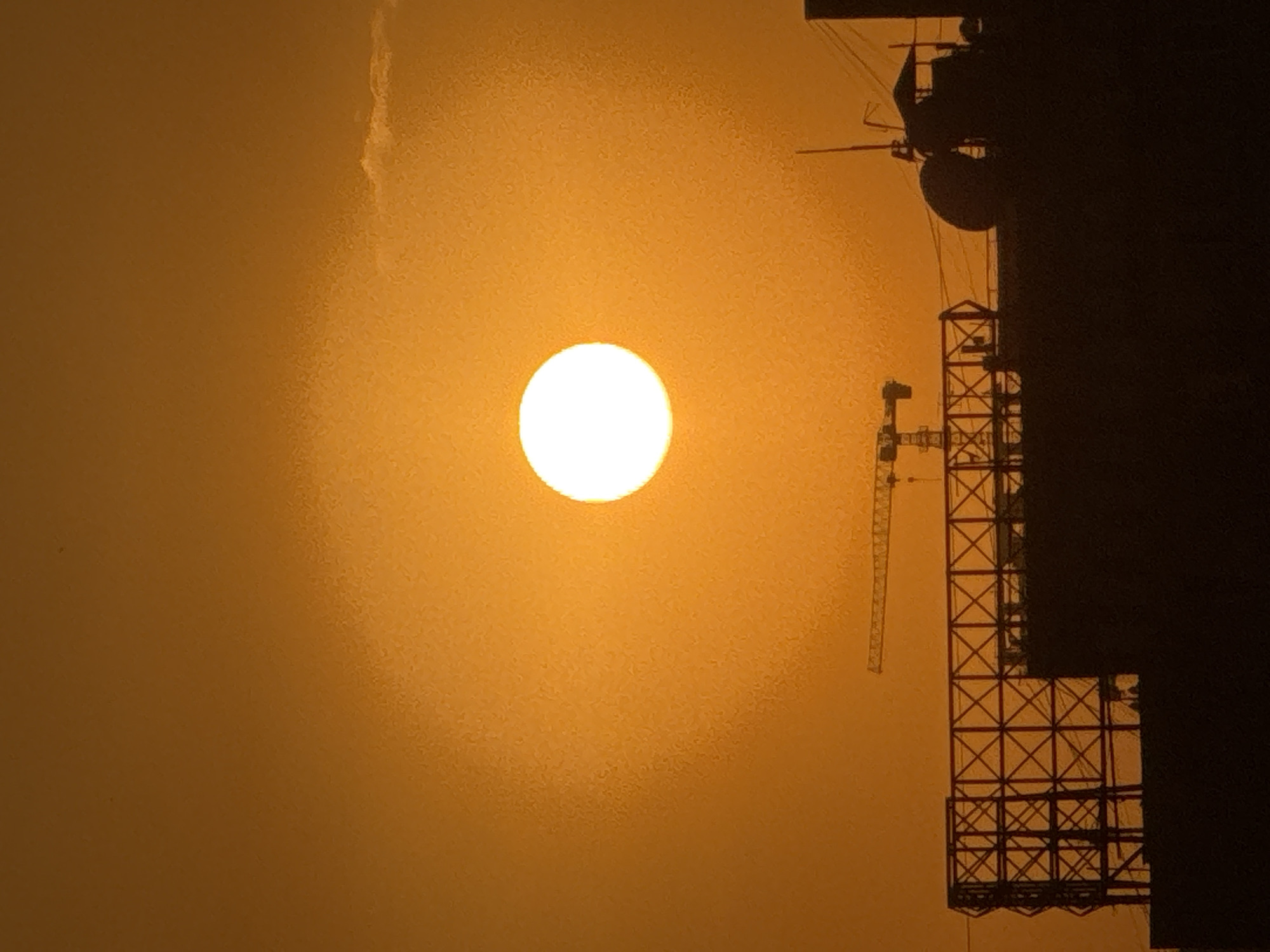 The image depicts a sunset over the city of Bishkek, Kyrgyzstan. A large, bright sun dominates the upper center of the frame, casting an orange glow across the sky. In the foreground, silhouetted against the bright sky, are the dark outlines of buildings under construction. These structures include a large, lattice-like framework, possibly part of a billboard or advertising structure, and a darker, more solid building with a roofed structure and antennas. A construction crane is visible in the mid-ground, also silhouetted. The overall scene suggests an urban environment undergoing development, set against the dramatic backdrop of a setting sun.