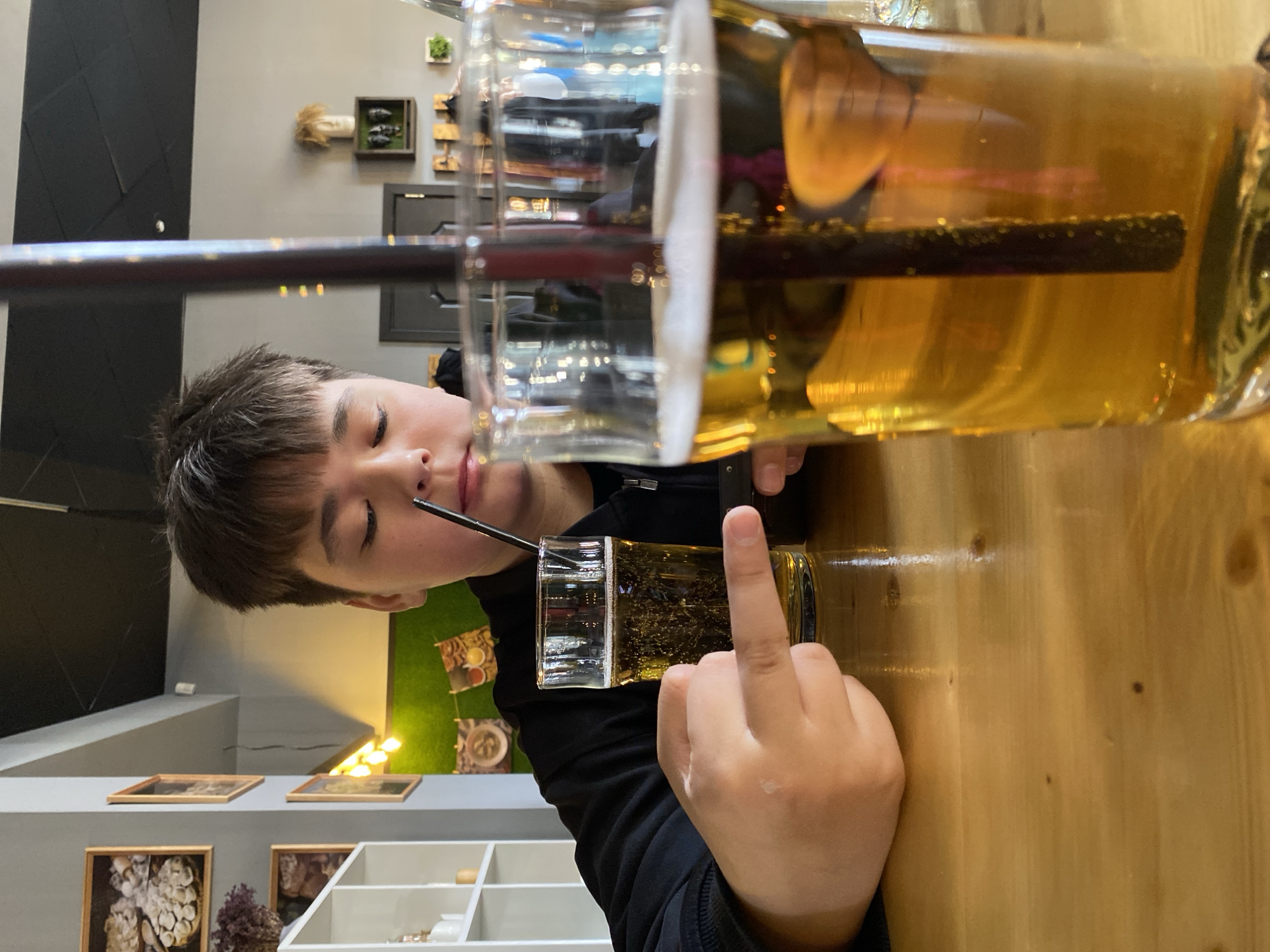 A young person, likely a boy, with short dark hair, is resting their head on or near a light wooden table in an indoor setting, probably a cafe in Votkinsk, Russia. Their eyes are closed or looking downwards, suggesting a moment of rest or contemplation.
In the foreground, a large, clear plastic cup or pitcher holds an amber-colored beverage with a dark straw and some visible particles, possibly tea or juice. A smaller, matching glass containing a similar drink is placed directly in front of the person. The person's hand rests on the table next to the smaller glass, with the middle finger extended upwards.
The background features contemporary decor, including a dark gray wall with a geometric black panel, and a lighter gray wall adorned with various framed artworks, decorative shelves, and small plants. A section of the wall is covered with artificial green turf-like material and illuminated by yellow lights. The lighting is bright and even, indicating an indoor environment that is well-lit. There is no visible text in the image.