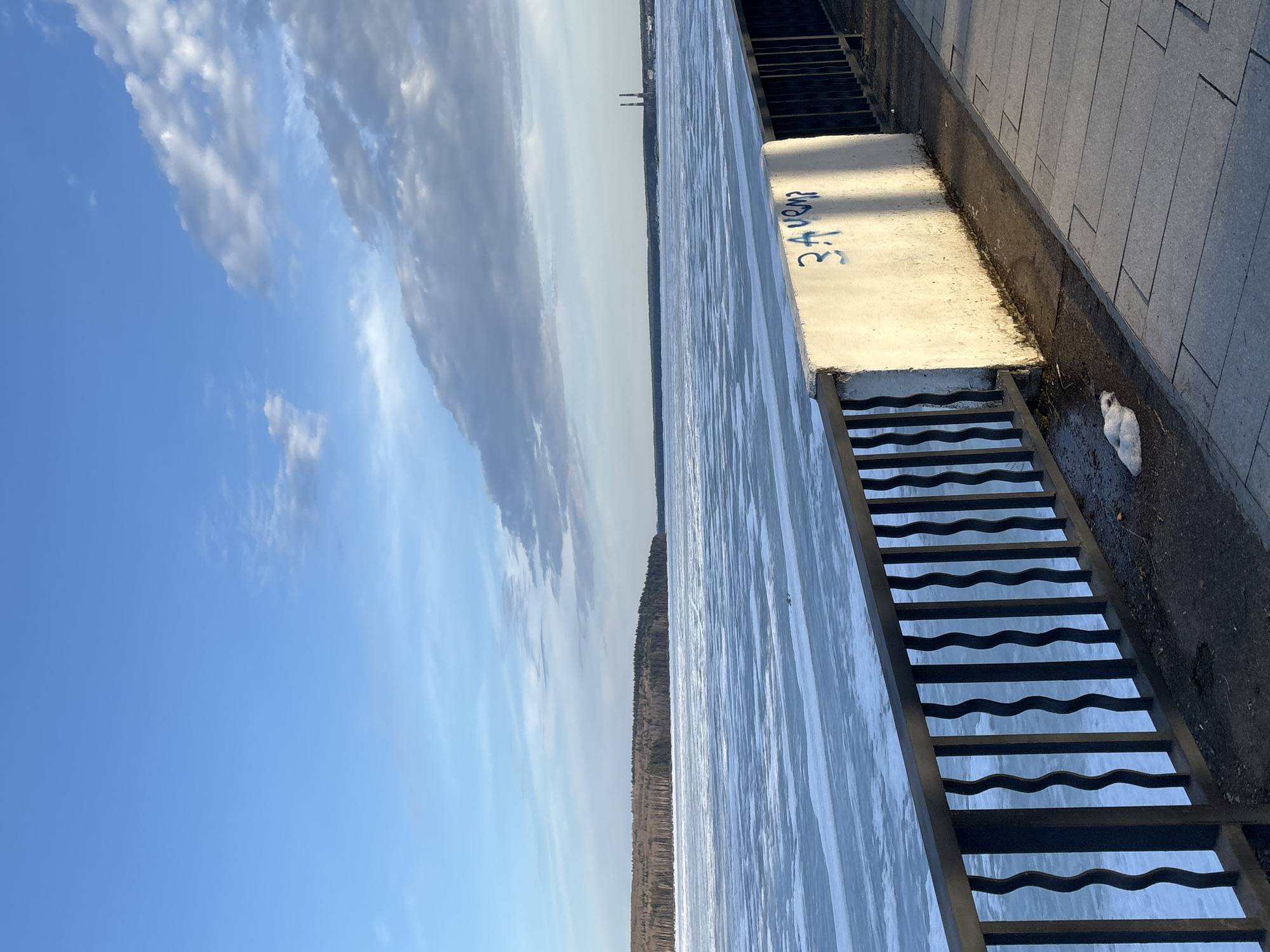 The image depicts a frozen body of water, likely a river or lake, in Chaikovsky, Russia. A set of metal stairs descends from a concrete embankment into the ice. Graffiti is visible on the concrete. Adjacent to the embankment is a paved walkway. The sky is blue with scattered clouds, and trees line the opposite shore of the water. In the distance, industrial structures are faintly visible. A piece of white trash is visible near the edge of the walkway.