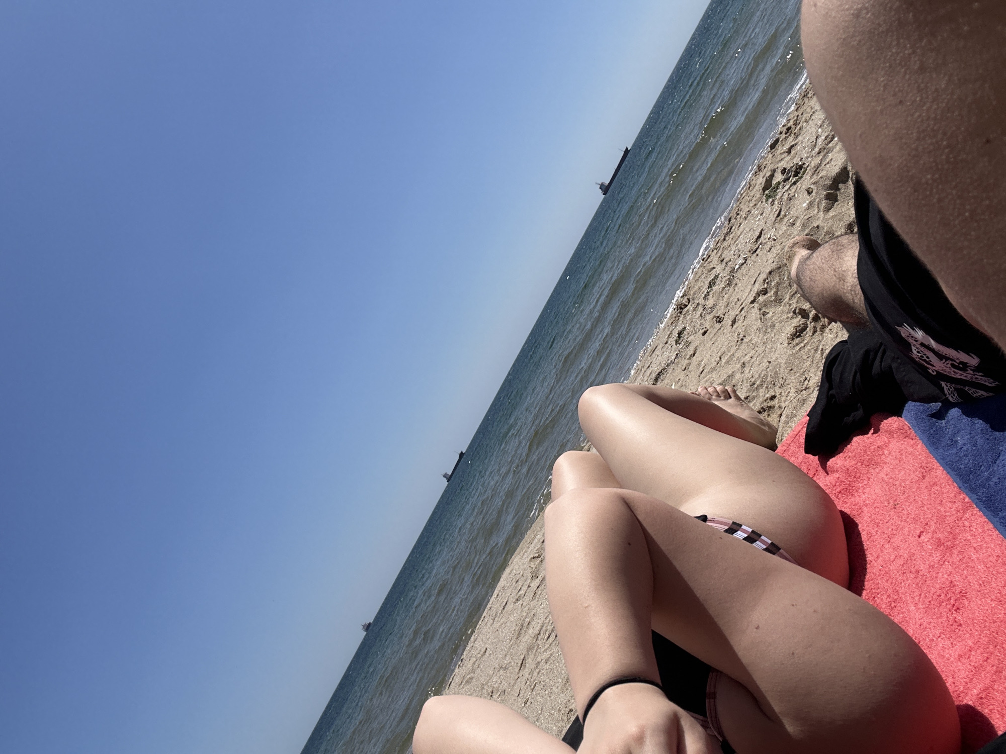 The image shows a sunny beach scene in Varna, Bulgaria. The view is from a low angle, looking toward the sea. The sky is clear blue. Several ships are visible on the horizon. The sandy beach is light tan and slopes down to the water. In the foreground, a portion of a person's legs and torso is visible on a red and blue towel. Another person's leg is visible in the right corner of the image.