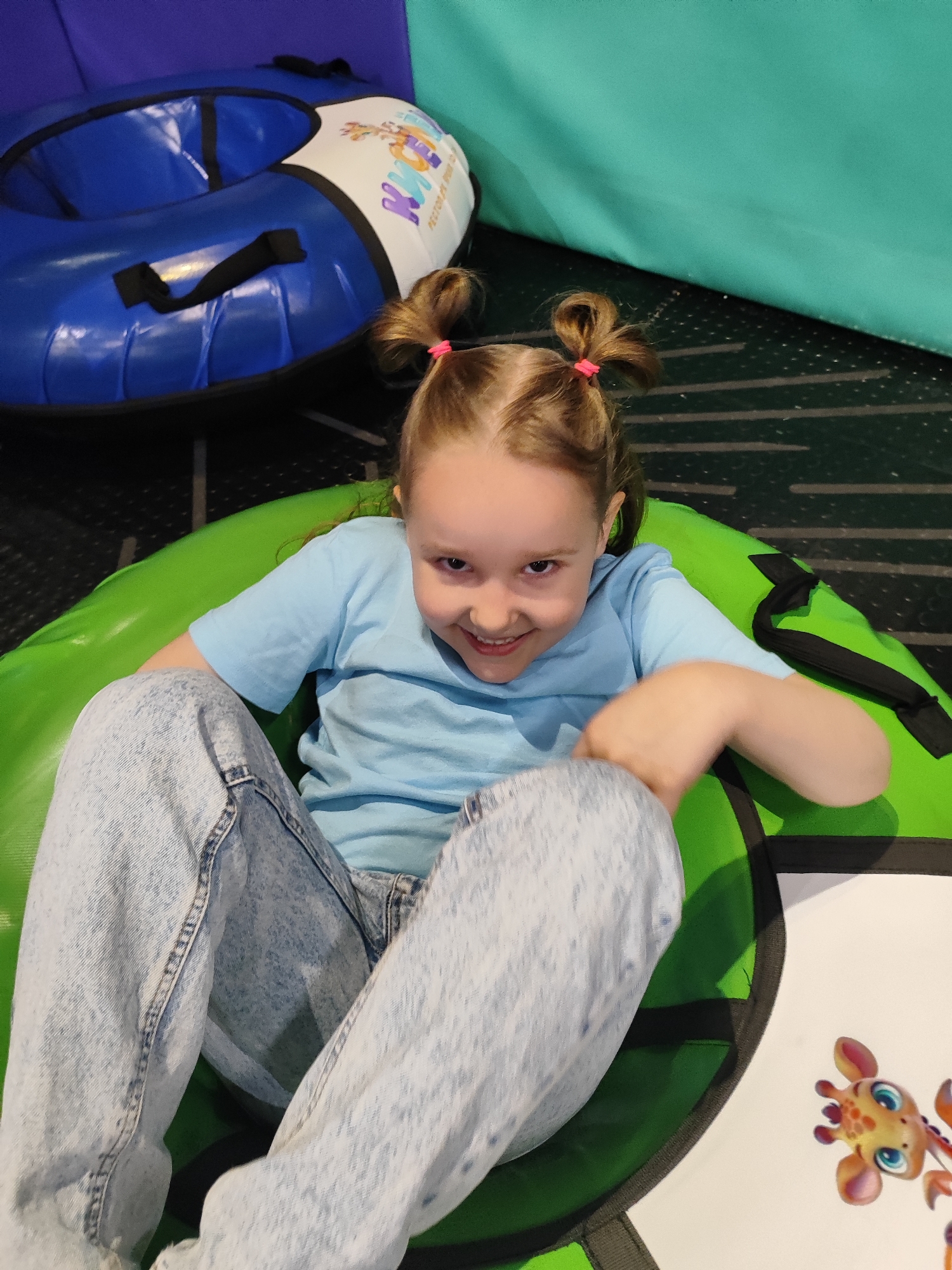 A young girl with light brown hair styled in pigtails secured with pink elastics, wearing a light blue t-shirt and acid wash jeans, is sitting in a green inflatable tube. The photo appears to be taken from above, showing the girl smiling at the camera. In the background, there is another blue inflatable tube and a wall with alternating teal and purple sections. The flooring is a dark, speckled surface. Part of a white sheet with a cartoon giraffe on it can be seen near the green tube. The location may be a recreational or entertainment center based on the presence of the inflatable tubes.
While I do not have any direct evidence the image was taken in the city of Krasnodar, Russia, it might be possible that I can find out the details with the help of other open-source tools and methods.