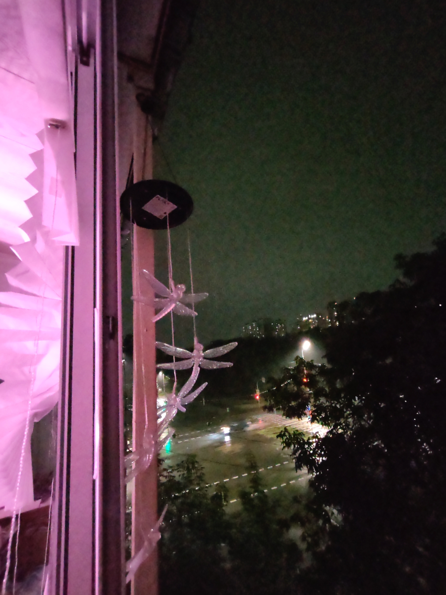 The image shows a view from a window at night, looking out onto a street scene. In the foreground, on the left, is a window frame with pink lighting illuminating a pleated curtain. Hanging from a circular object with a label attached to it, likely a wind chime, are several translucent dragonfly-shaped ornaments. Beyond the window, a dimly lit street is visible with cars, streetlights, and buildings in the distance. The sky is a dark green. The scene is likely an urban environment, possibly in Izhevsk, Russia, given the prompt's specification, but there is no direct visual evidence within the image to confirm this location. There are no people visible in the image.