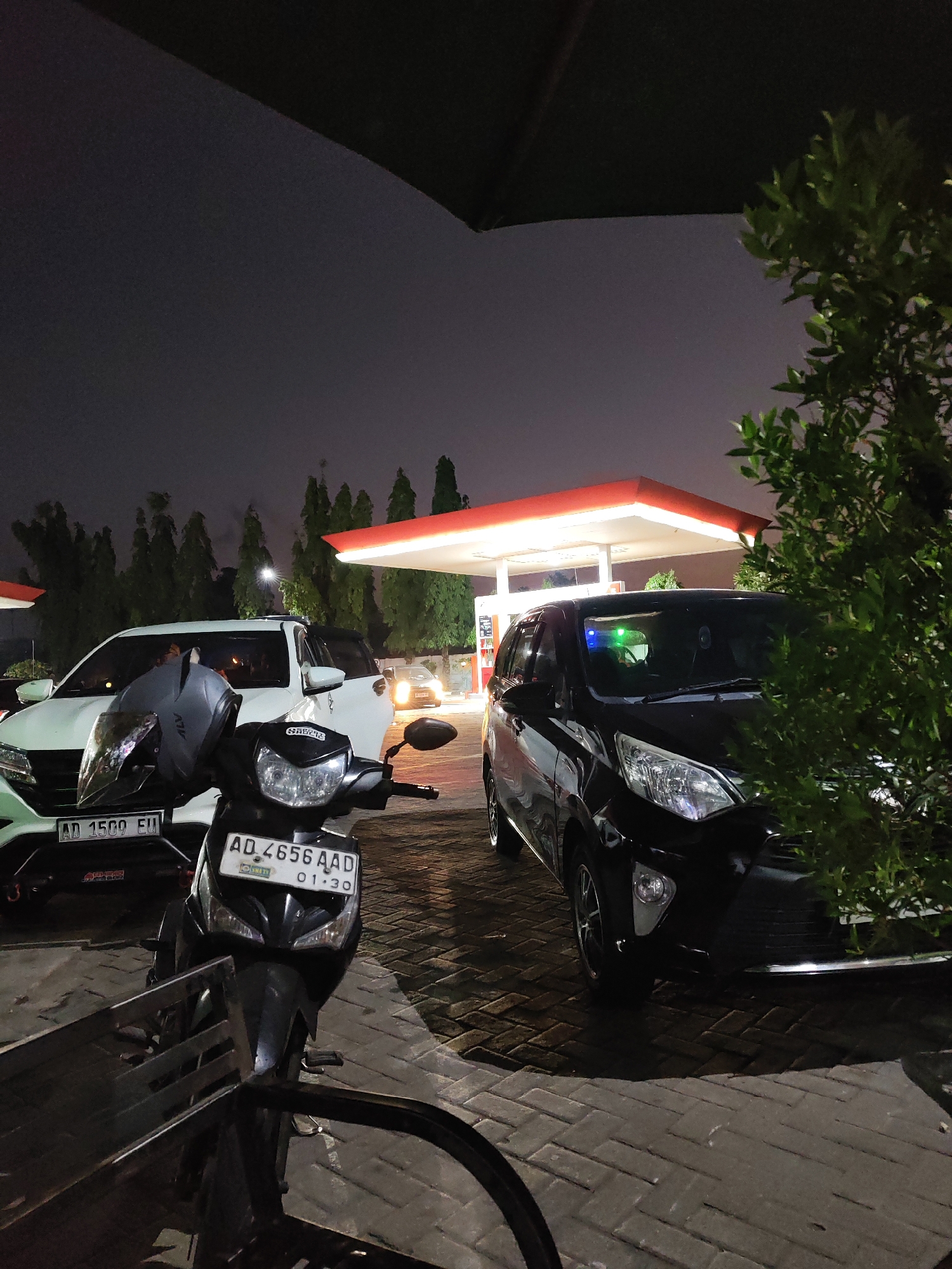 At night, a black car and a white SUV are parked at a gas station in Geneng, Indonesia. In the foreground, a motorcycle is parked, with a helmet on its handlebars. A leafy green bush is to the right of the black car. The gas station canopy is illuminated with red and white lighting. The sky is dark and overcast.