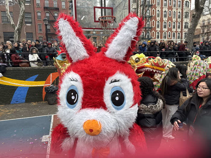 A large, red and white bunny costume with large eyes is prominently featured in the foreground. Behind the bunny, a traditional Chinese dragon dance is taking place in what appears to be an outdoor park setting. A crowd of spectators is gathered on an elevated concrete barrier, watching the festivities. The backdrop includes brick buildings and trees, suggesting an urban environment. The image captures a vibrant cultural celebration.