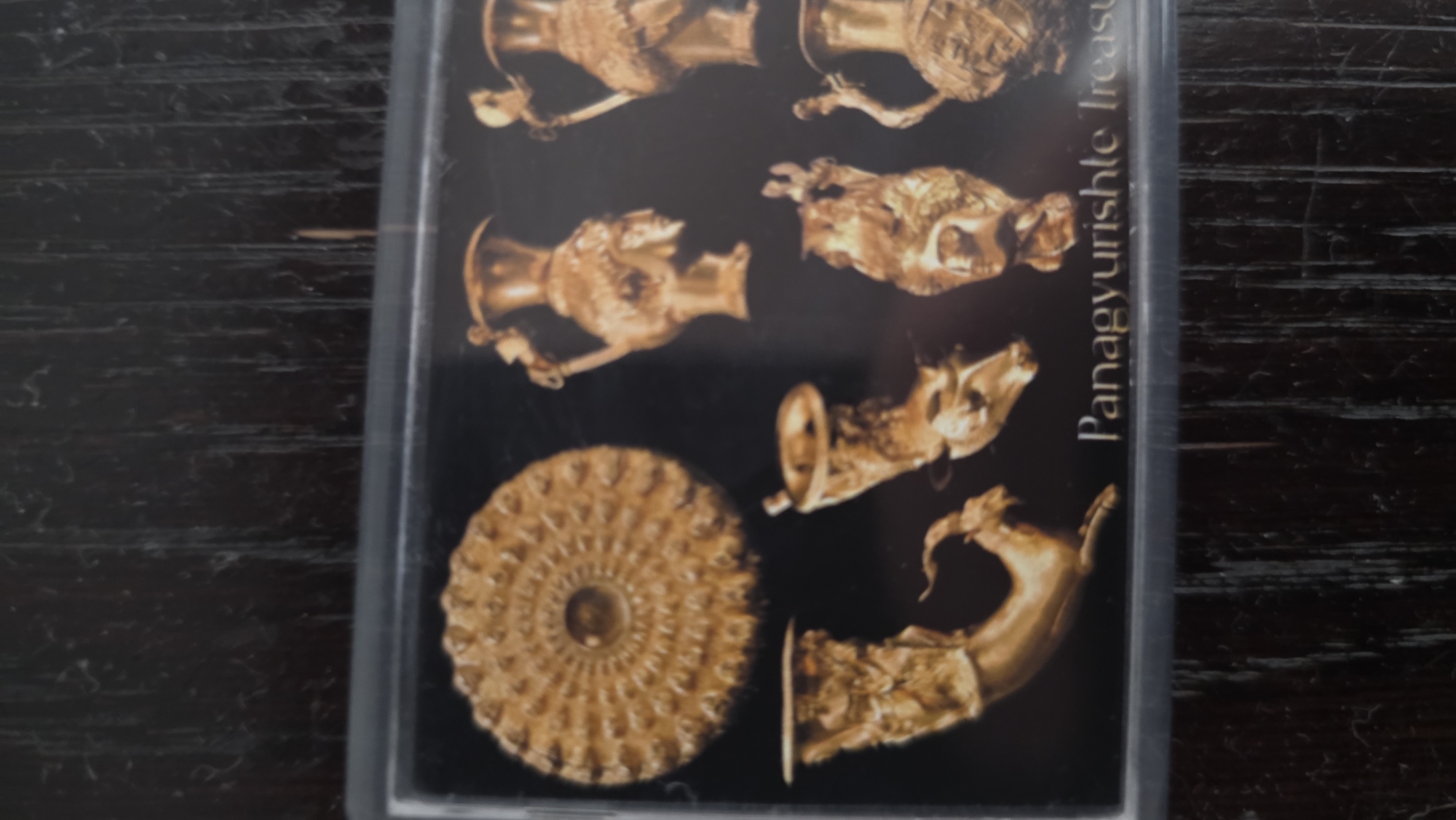 The image depicts a close-up of a card or photograph showing images of golden artifacts against a black background. Text reading "Panagyurishte Treasure" is visible on the right side of the image, oriented vertically. The artifacts are ornate, likely made of gold, and include vessels with animal-shaped handles and a decorative circular element.
The Panagyurishte Treasure is a Thracian gold treasure discovered in Panagyurishte, Bulgaria, which is located in the Pazardzhik Province and not directly in Blagoevgrad. The artifacts are on display in Bulgaria's National History Museum in Sofia, not in Blagoevgrad.