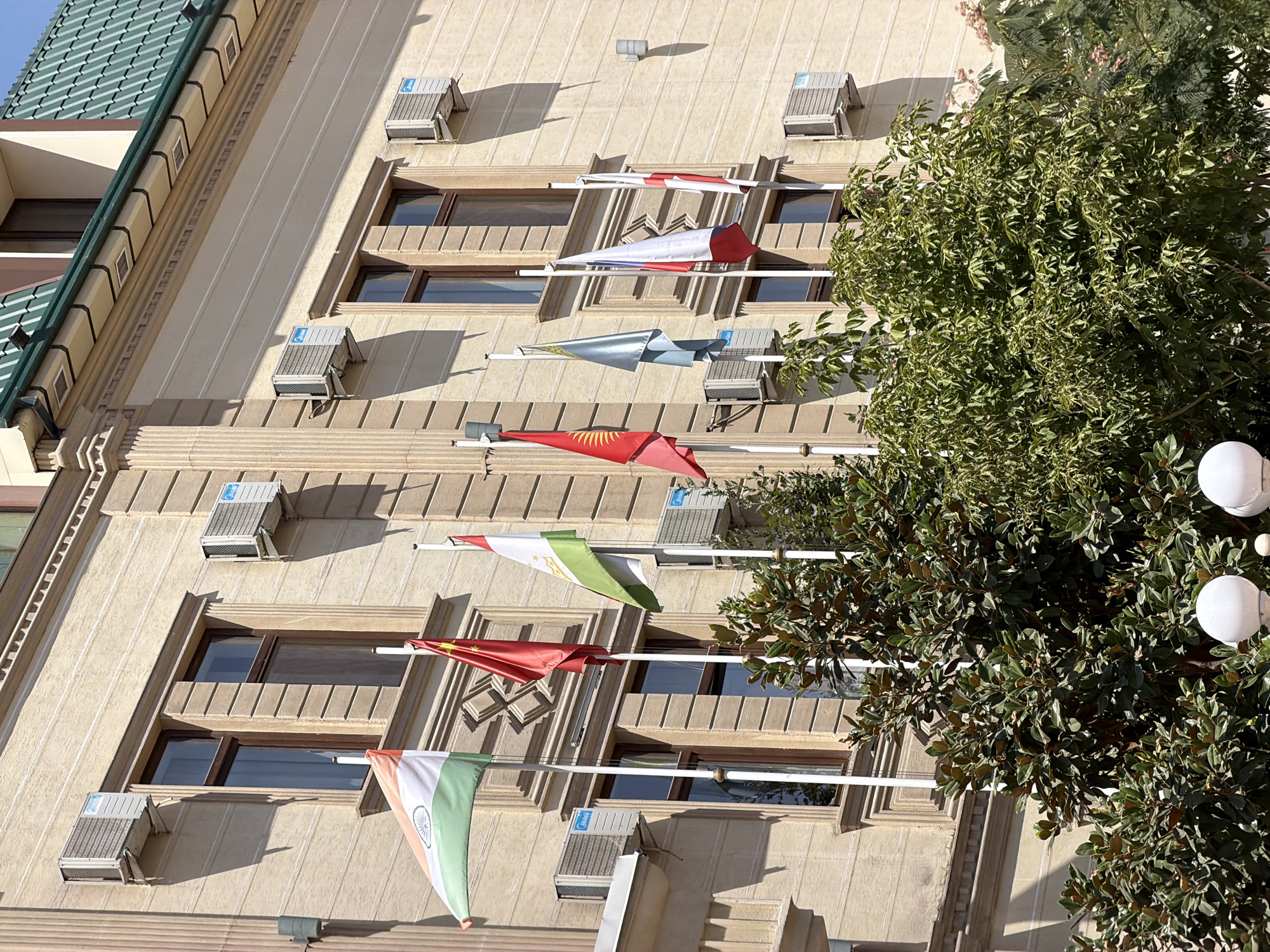 The image shows the facade of a building in Tashkent, Uzbekistan, adorned with several flags. The building is light beige with ornate architectural details and features multiple windows, some with dark wood frames. Air conditioning units are visible mounted on the exterior walls. In front of the building, a row of flags on poles is displayed, including the flags of India, China, Tajikistan, Kyrgyzstan, Kazakhstan, and possibly others. Lush green trees and bushes are in the foreground, partially obscuring the lower part of the building. The scene appears to be a sunny day.