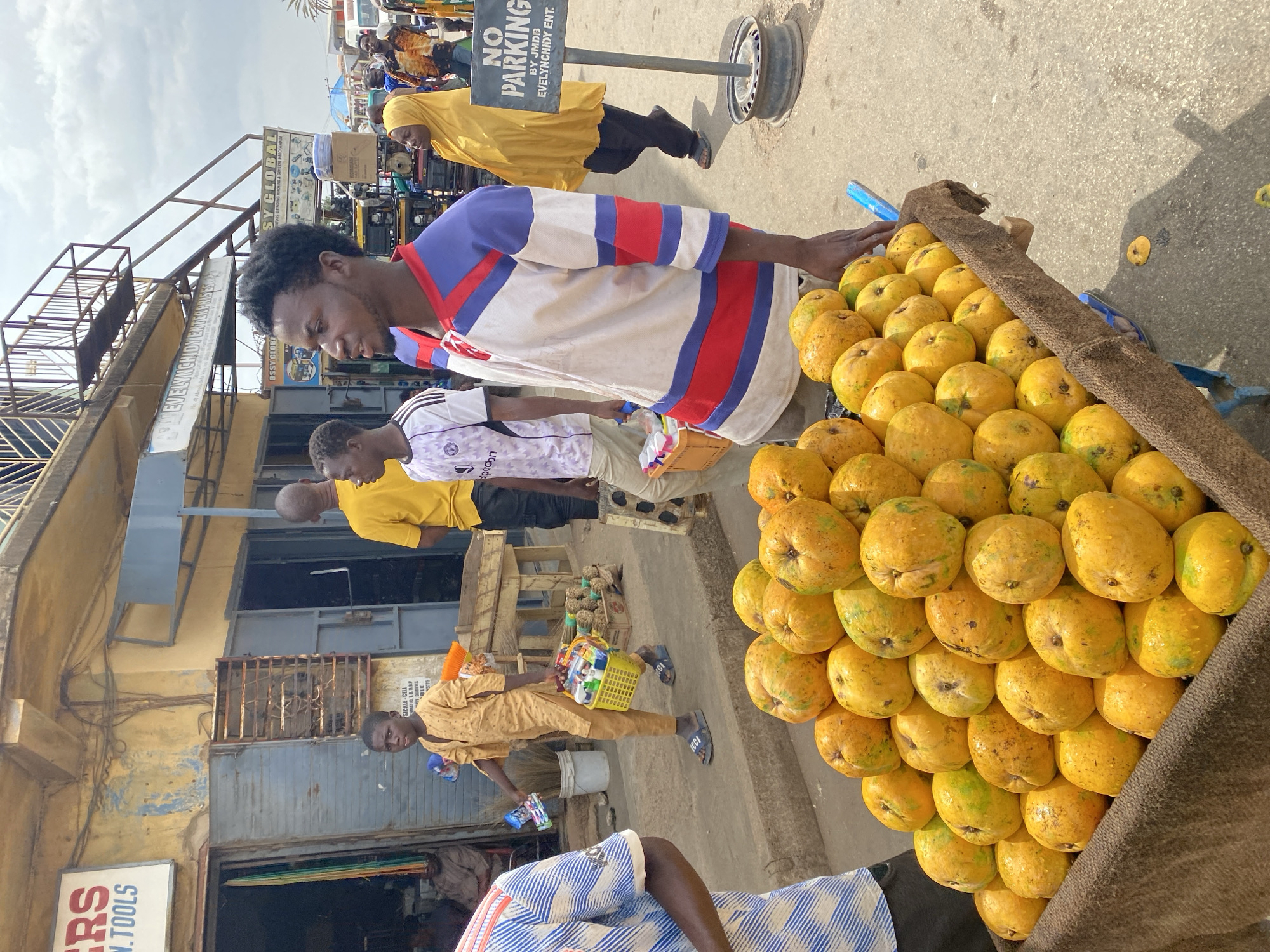 A street scene in Jos, Nigeria, shows a man in a white, red, and blue striped shirt tending to a cart overloaded with yellow fruits, possibly mangoes or similar local produce. The fruits are stacked high and covered with a brown burlap-like material. The man is looking down at the cart. Several other people are present in the background, including a man in a yellow shirt and another in a patterned white shirt. To the left of the fruit cart, there are buildings with faded paint and various signs. One sign reads "EVELYNCHIDY ENTERPRISES", and another partially reads "---RS N.TOOLS". A "NO PARKING" sign is visible in the background, constructed of a metal pole topped with a car wheel. The street appears to be in a busy commercial area.