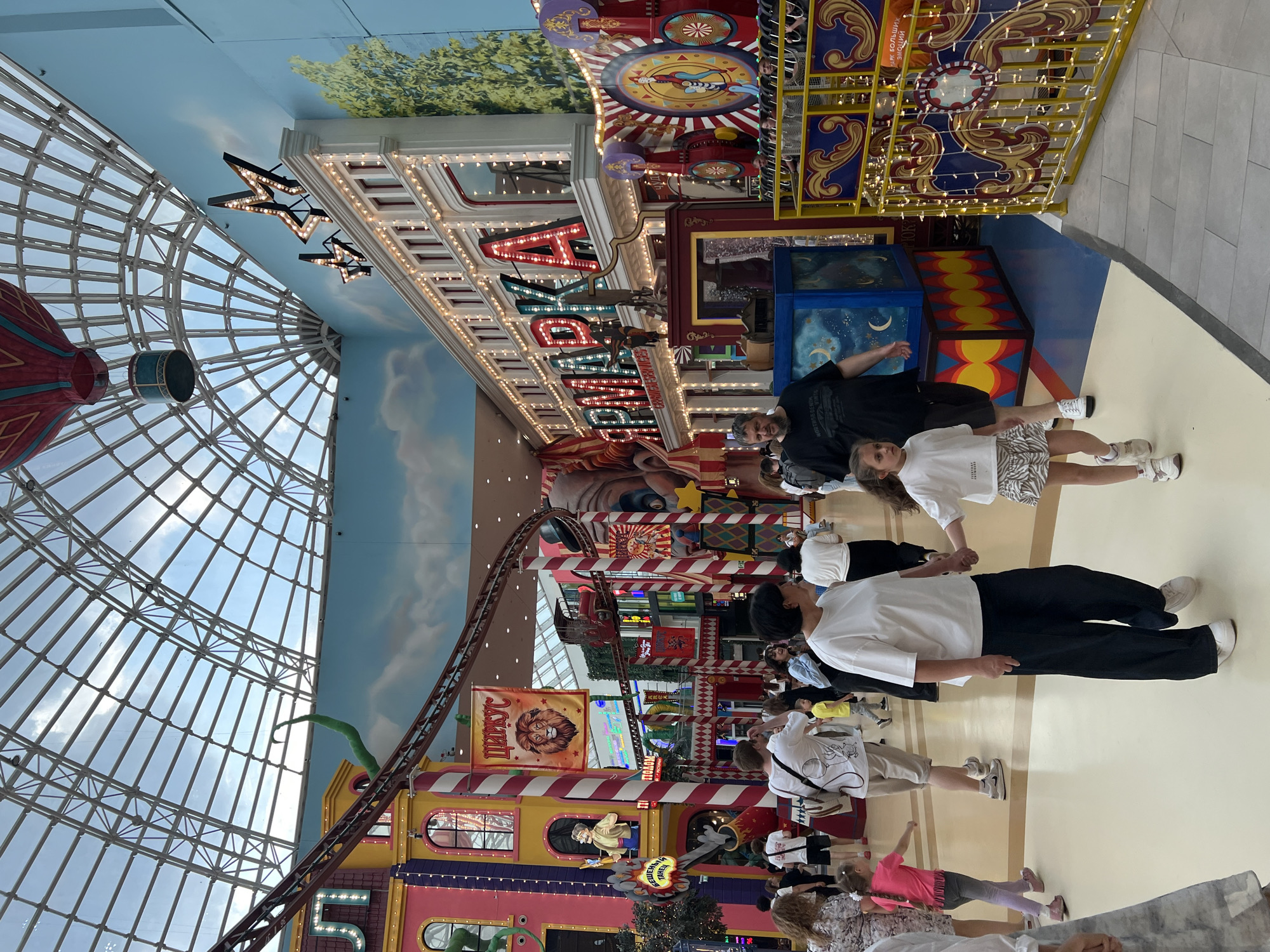 The image depicts the interior of a brightly colored amusement park or entertainment complex, likely located in Moscow, Russia. A large, transparent, curved roof structure allows natural light to filter in, illuminating the scene. In the background, a roller coaster track winds through the space, above various themed structures that resemble buildings with circus-like decorations. These structures are painted in vibrant colors such as pink, yellow, and blue, and feature whimsical elements like large cartoon characters and illuminated signage. One prominent sign displays "ЯРМАРКА" in large, glowing letters, which translates to "Fair" or "Market" in Russian, suggesting a carnival or market-themed attraction.
The foreground and midground are populated by a diverse group of people walking and interacting. Many are dressed in casual attire, indicating a relaxed atmosphere. Families with children are visible, with parents holding hands with their children. The flooring appears to be a polished, light-colored material, with segmented sections in different colors, creating a dynamic visual effect. The overall ambiance is cheerful and energetic, typical of a family entertainment venue.